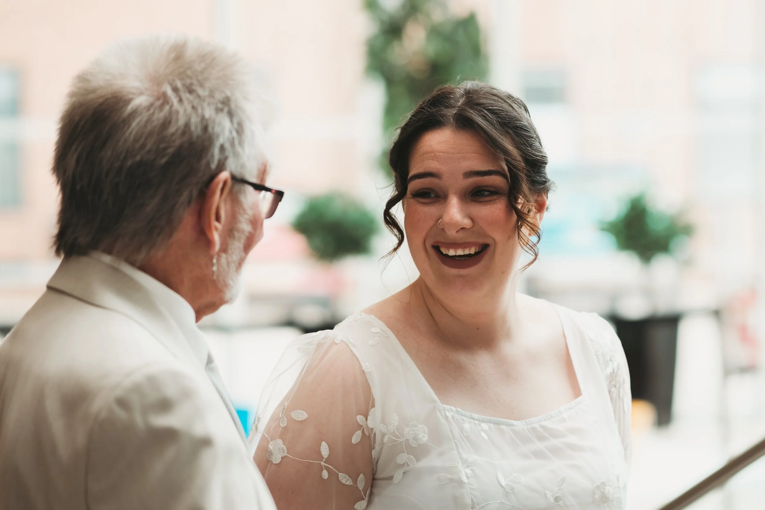 A young woman with dark curly hair and a nose ring smiling and talking to an older man with white hair, glasses, and a white jacket in an indoor setting with blurred background.