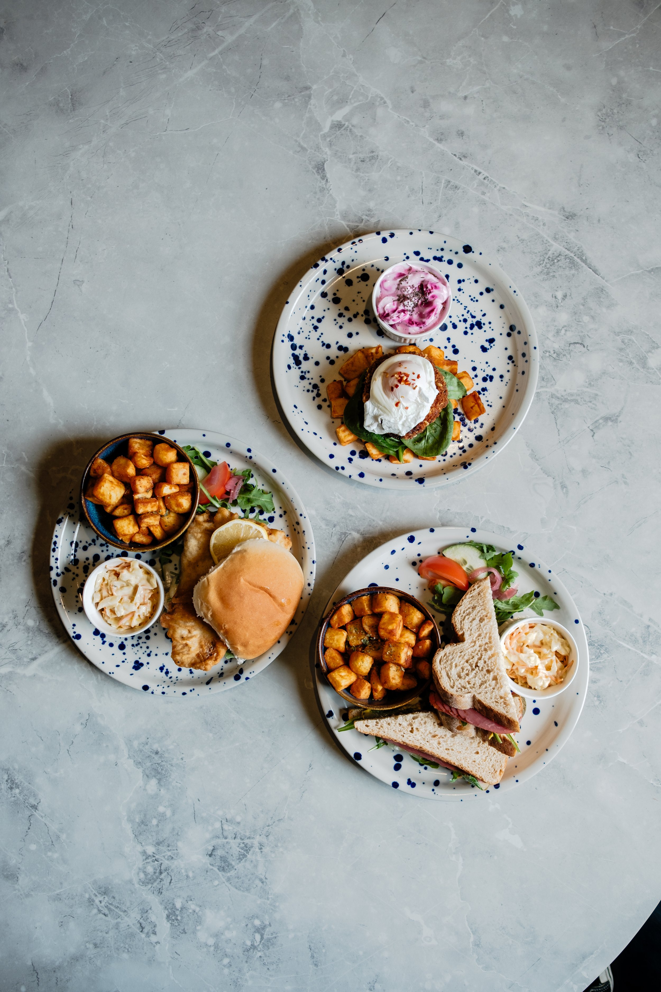 Three plates of food on a gray marble table, including sandwiches, salad, roasted potatoes, and a side of coleslaw.