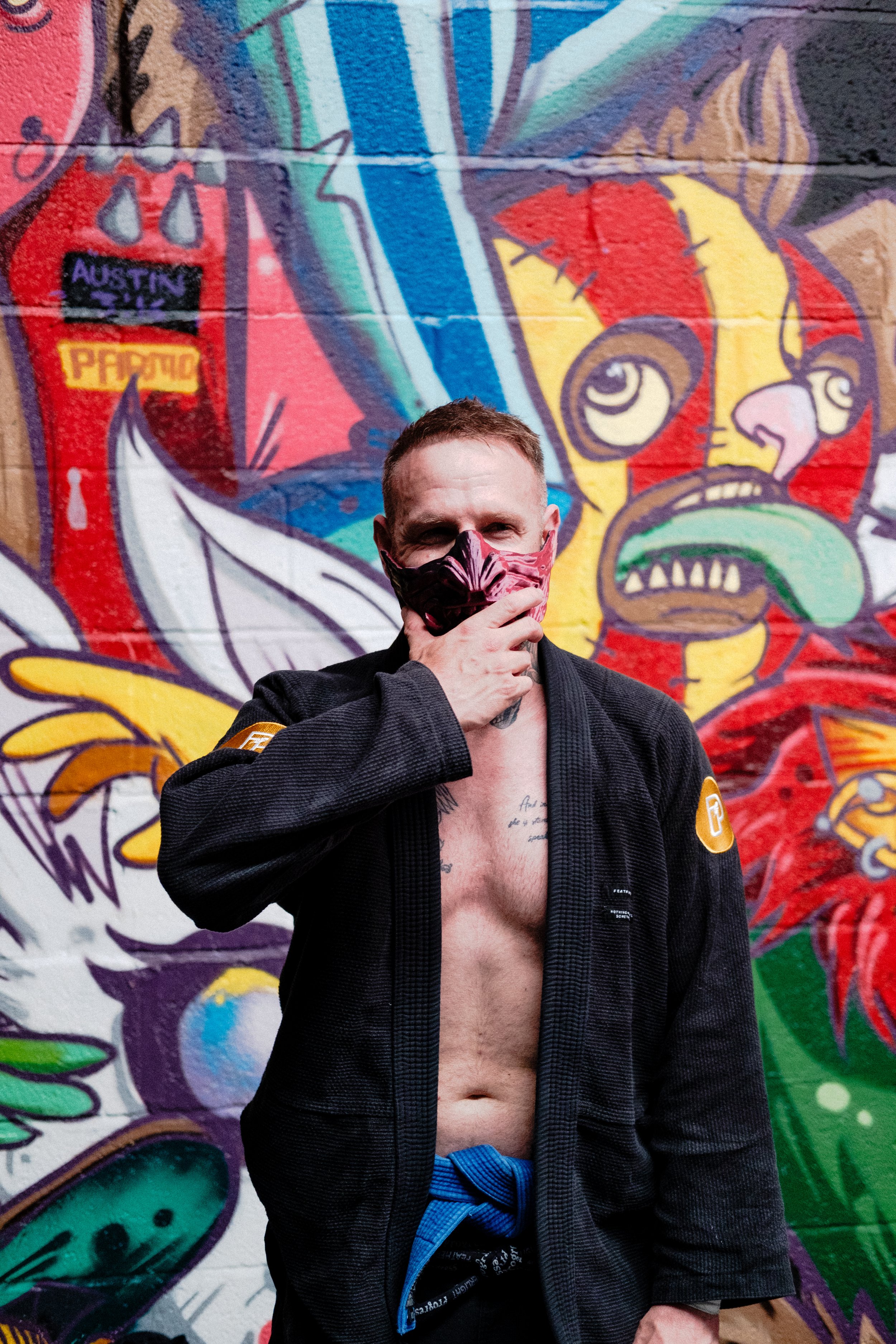 A man standing in front of a colorful graffiti mural, wearing an open martial arts gi with a blue belt, a face mask, and showing tattoos on his chest.