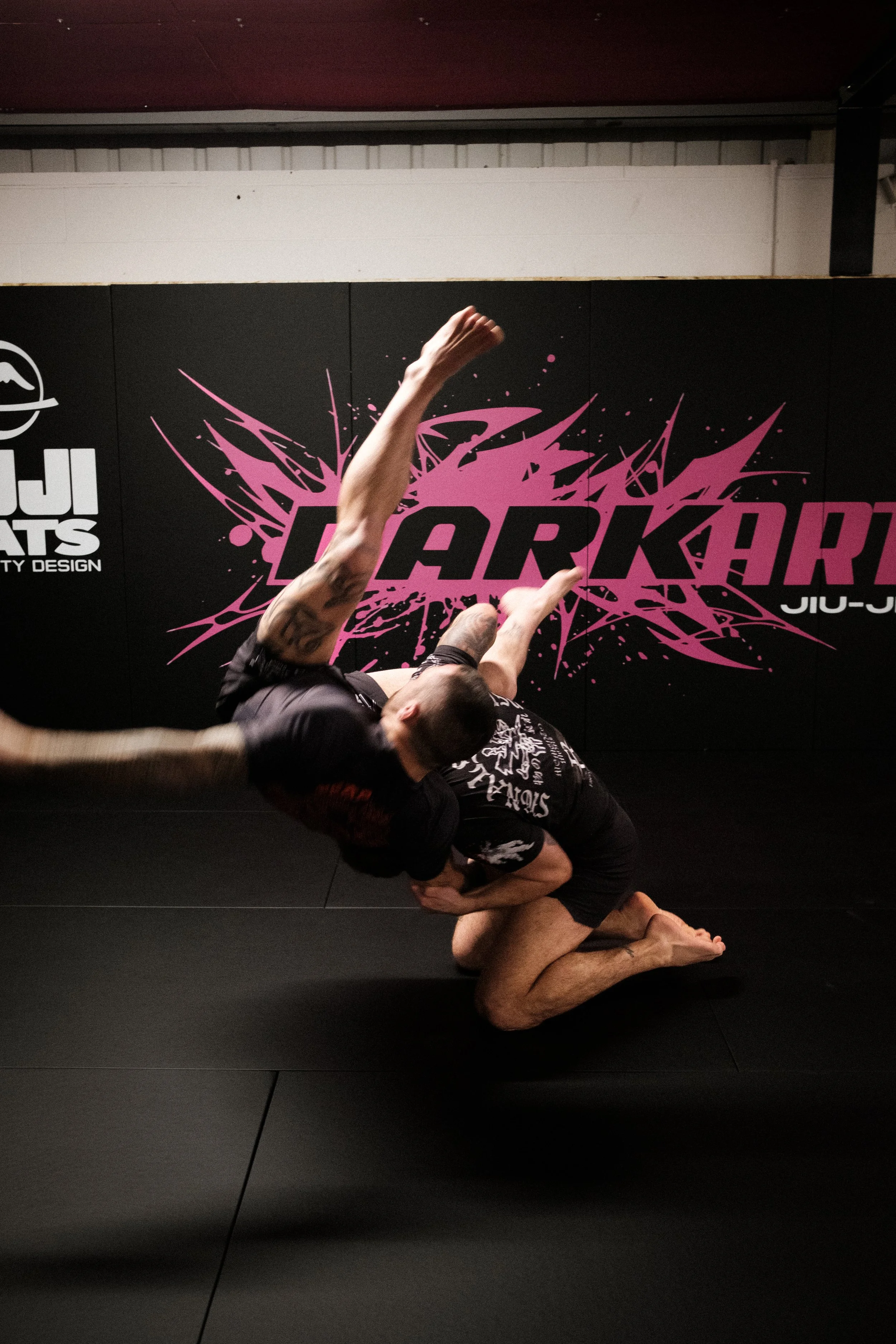 Two men practicing Brazilian Jiu-Jitsu on a black mat with a black wall in the background. One man is executing a submission move while the other is on his knees and leaning forward.