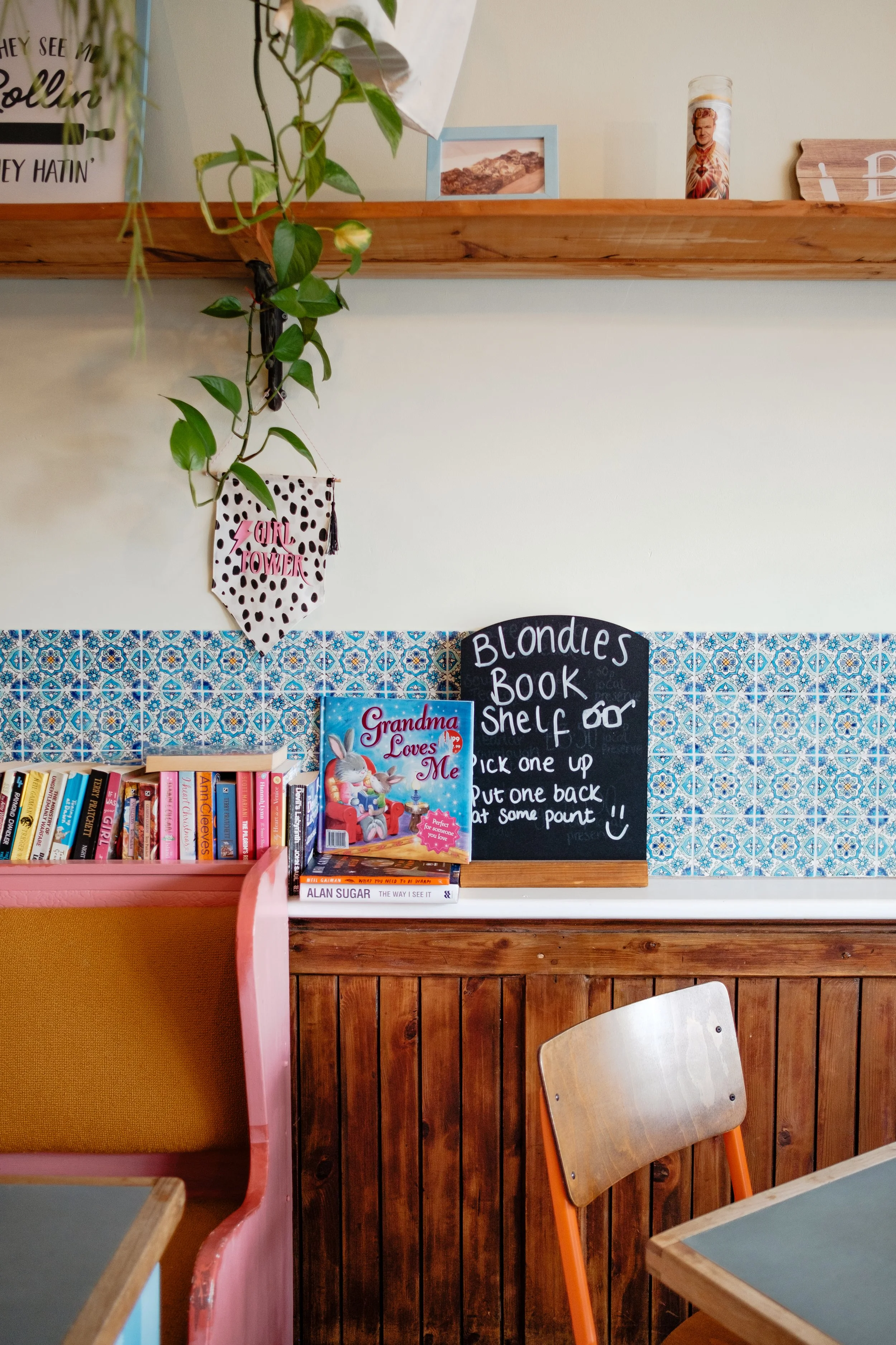 A bookshelf with colorful books next to a black chalkboard sign that says "Blondes Book Shelf" and provides instructions. A children's book titled "Grandma Loves Me" is visible on the shelf. There is a wooden chair with a light-colored seat and an or