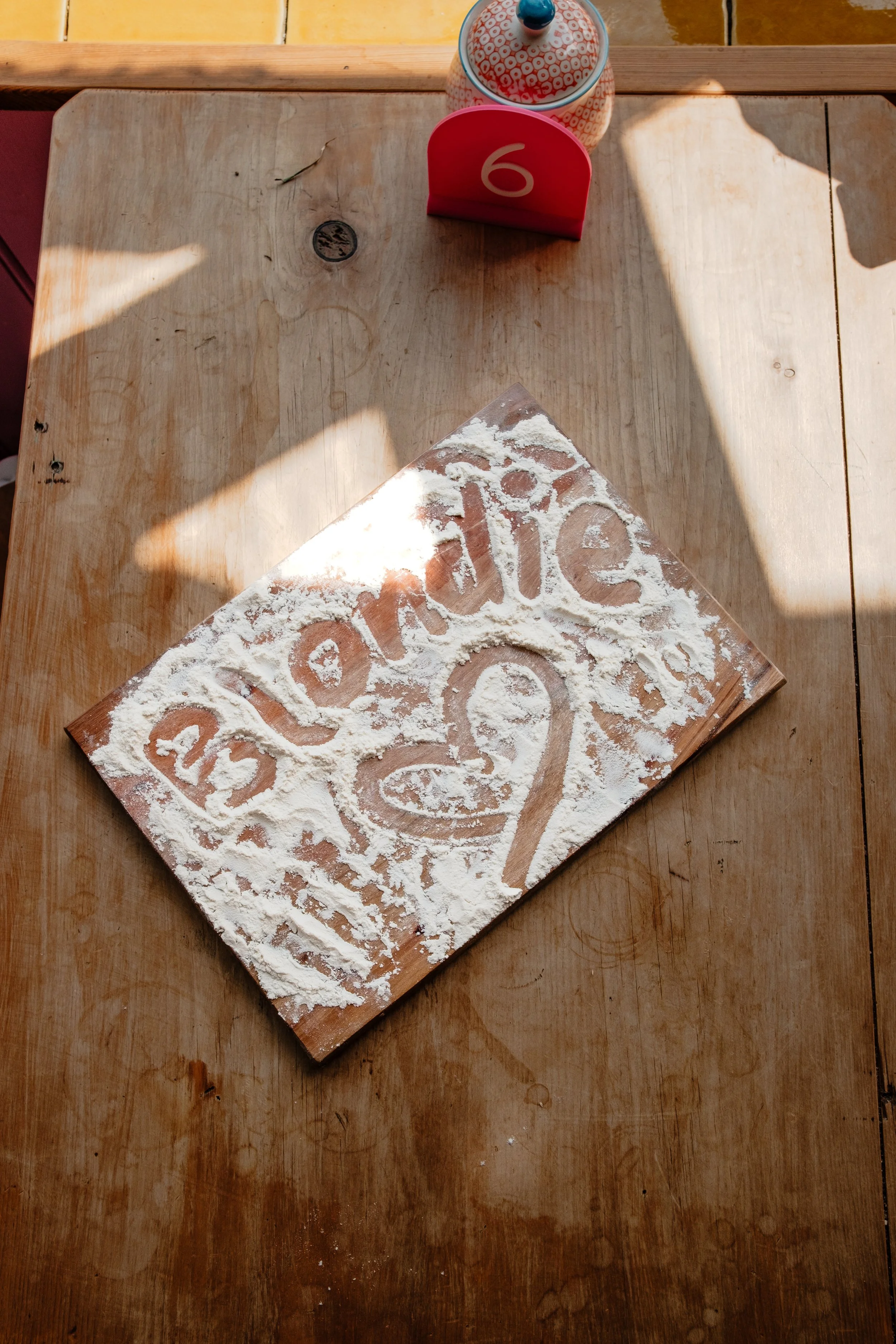 A wooden table with flour spread on it, forming the word 'Love'. A pink table sign with the number 6 is on the table. A ceramic jar with a lid and a colorful pattern is also present on the table.