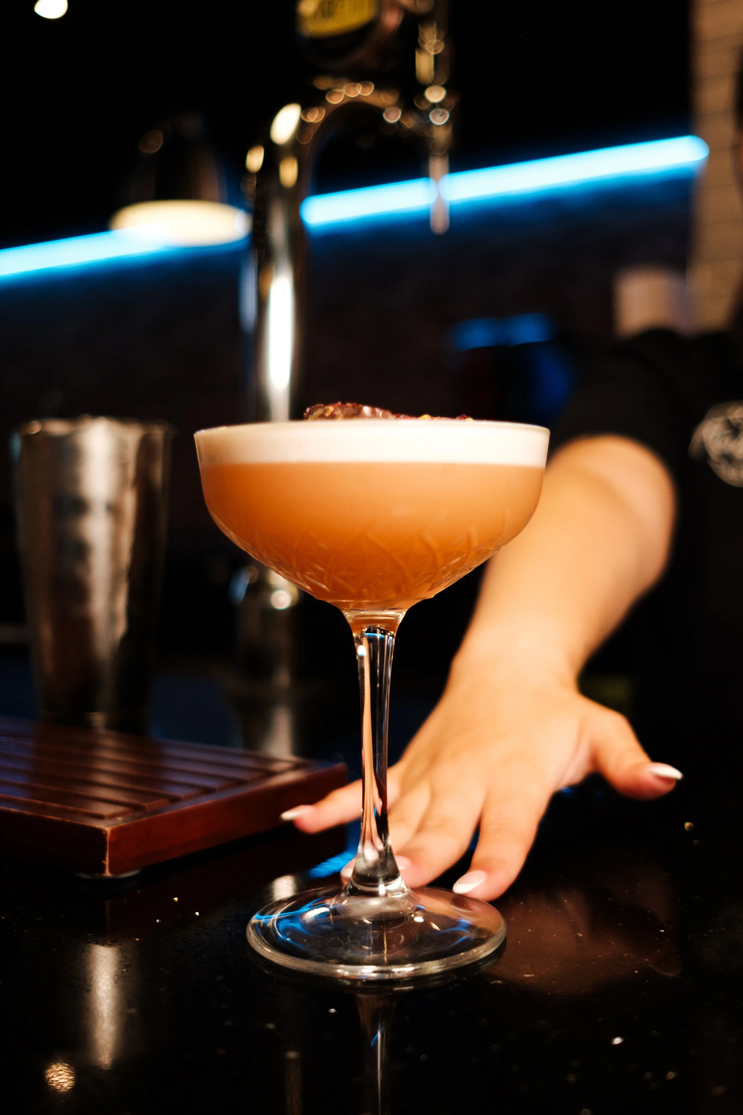 Close-up of a pink cocktail in a coupe glass with a frothy top, held by a person's hand, with a bar counter and a handrail with blue neon lights in the background.