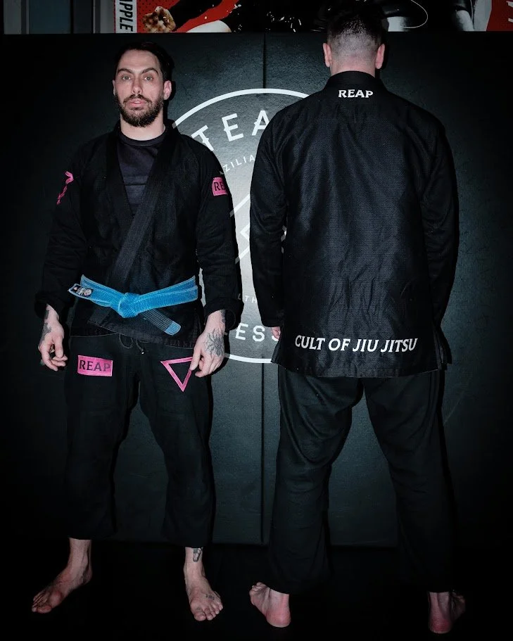 Two men in black martial arts uniforms standing against a black background, with one facing forward and the other with his back to the camera. The uniforms have patches and text, including 'REAP' and 'CULT OF JIU JITSU'.