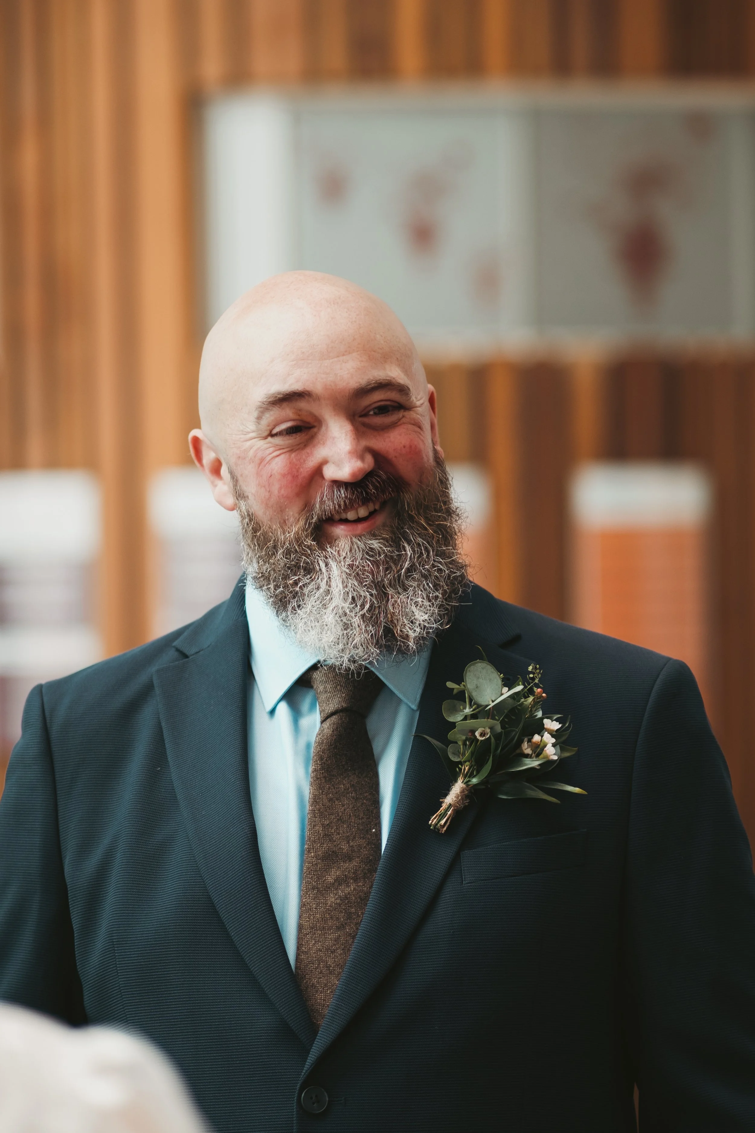 A smiling bald man with a beard dressed in a dark suit, light blue shirt, and brown tie, wearing a boutonniere with greenery and small flowers, standing indoors with wooden paneling and cubical storage units in the background.