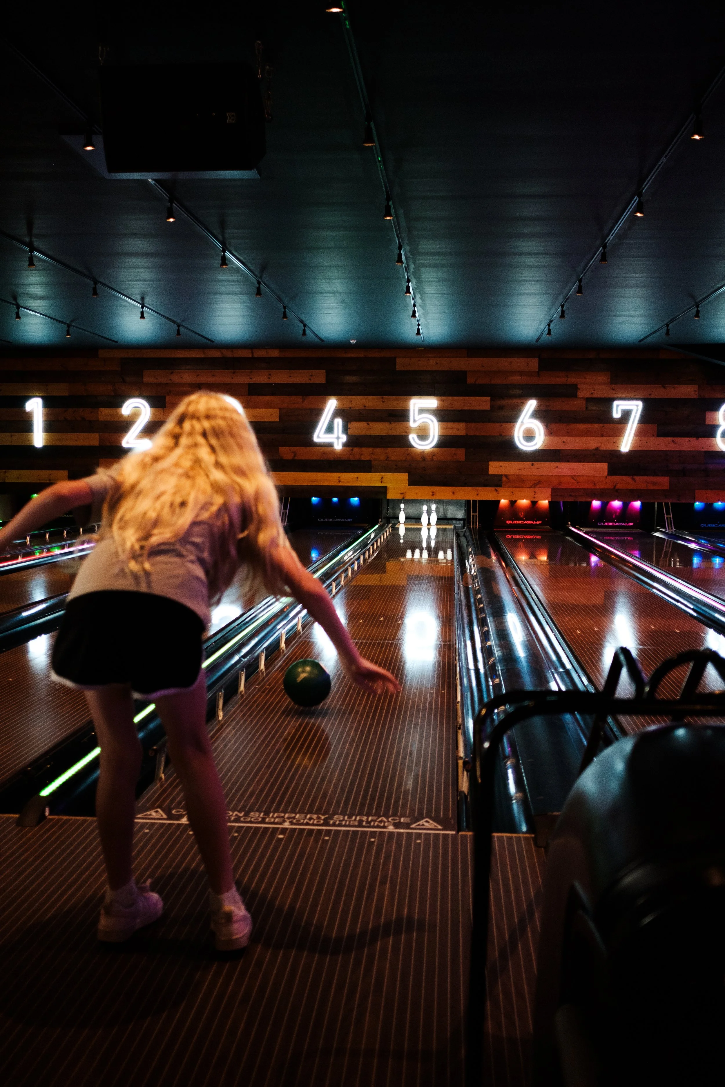 A girl bowling at an indoor alley, with glowing pins at the end of the lane and large illuminated numbers on a wooden wall behind. She is about to release a green bowling ball.