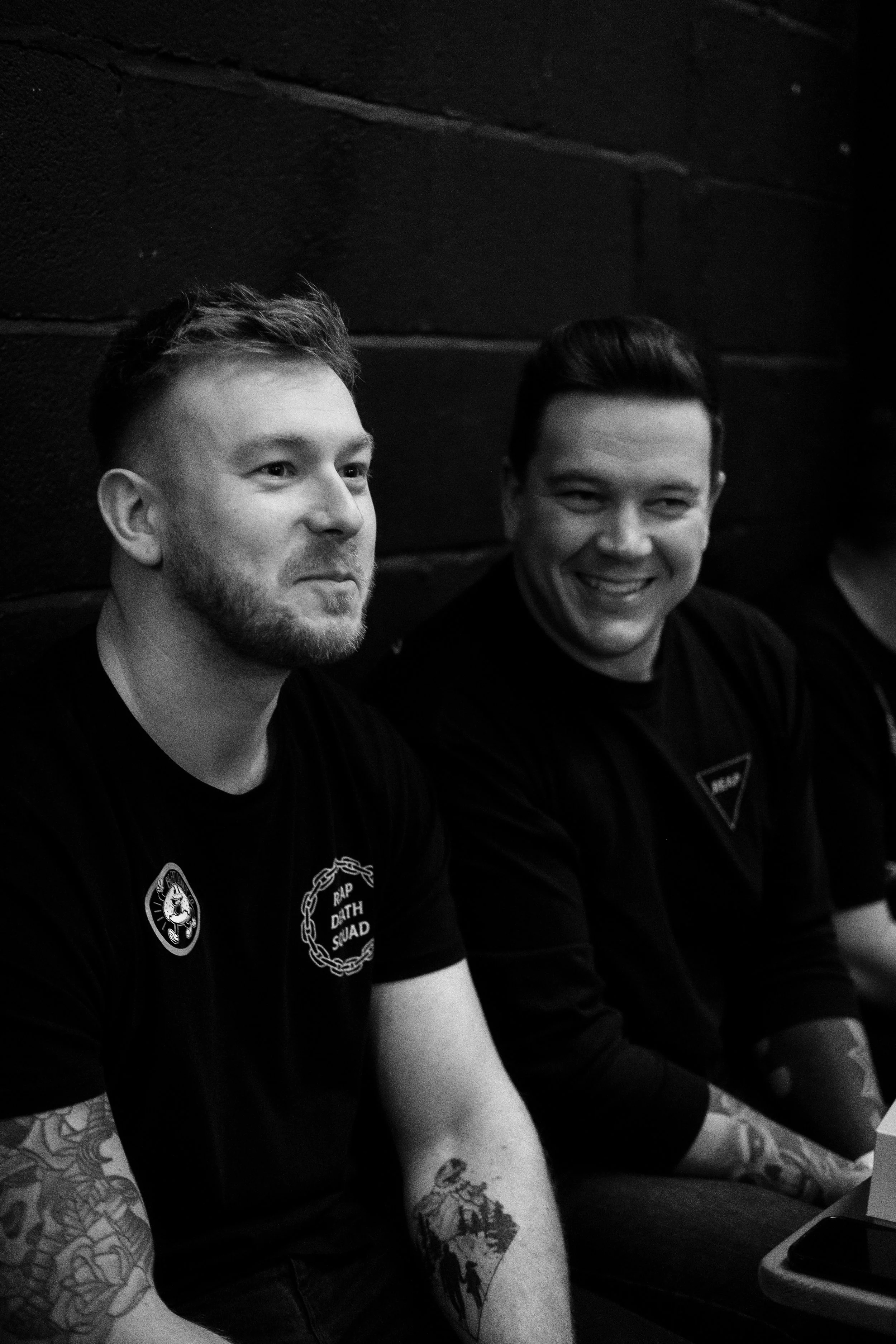 Two young men sitting against a dark wall, smiling and looking to the side, wearing dark T-shirts with patches and tattoos on their arms.