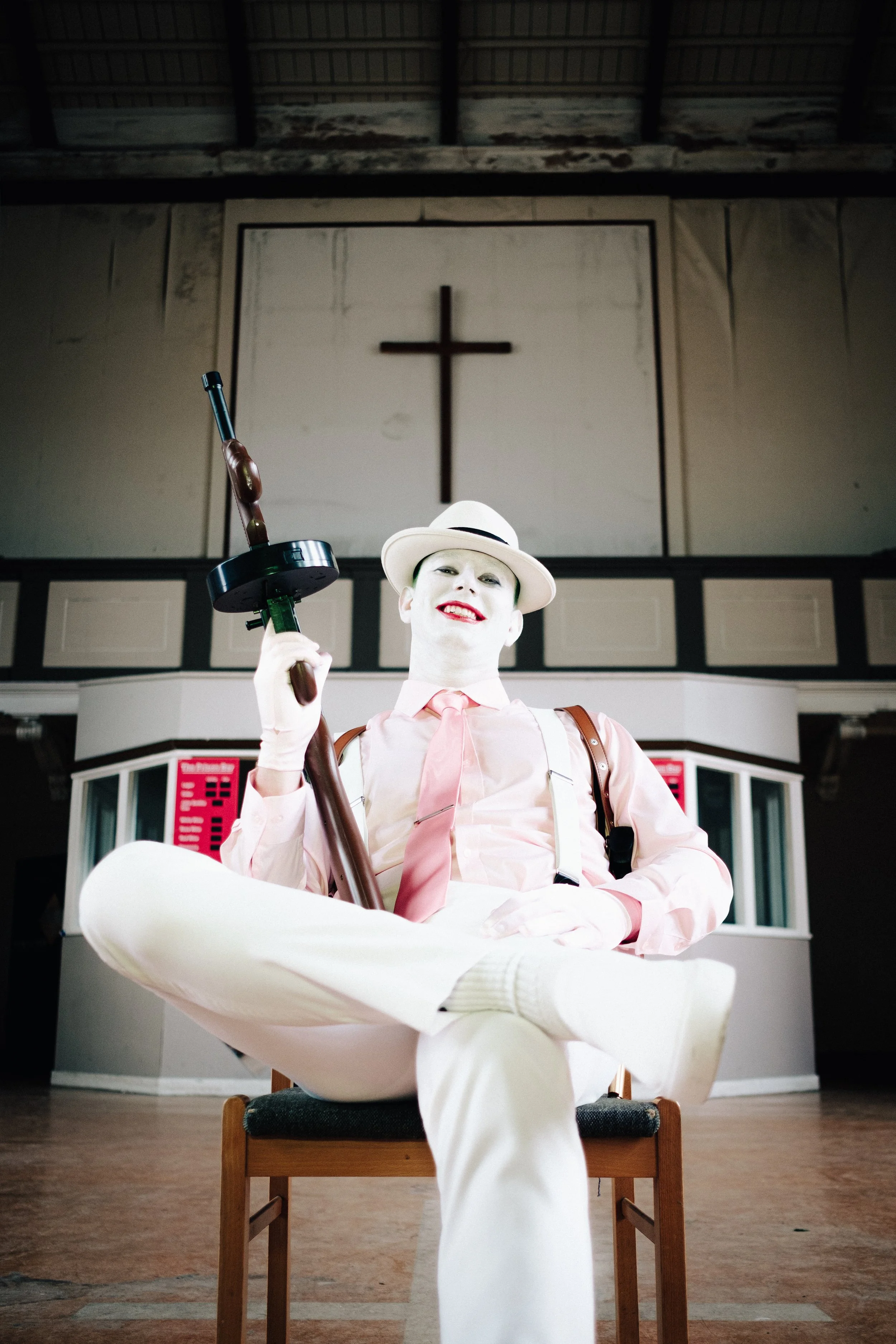 A person wearing white makeup, a white hat, and pastel-colored clothes sits on a chair inside a church. They hold a prop gun with a wooden handle. The background features a large cross and church interior.