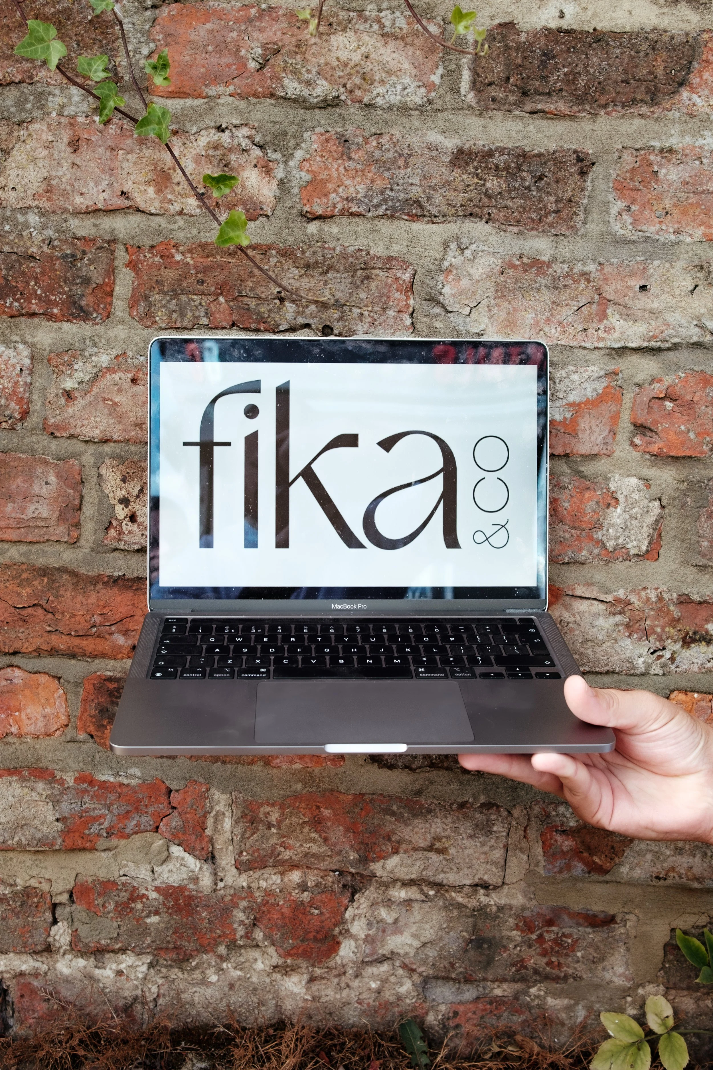 A person holding a MacBook Pro laptop with a screen displaying the word 'fika' against a brick wall background.