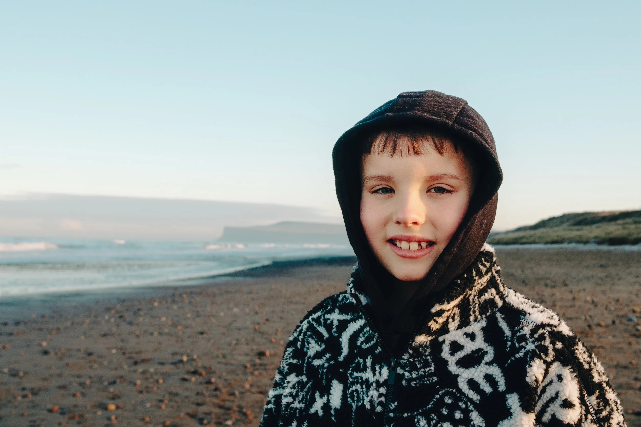 A young boy with blond hair and blue eyes smiling on a beach during sunset, wearing a black hoodie and a patterned black and white jacket.