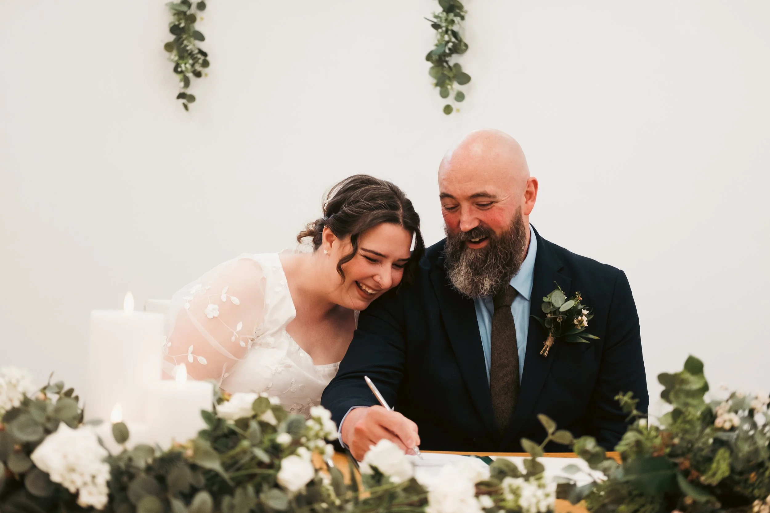 A bride and groom are sitting at a table, smiling and signing a document, likely during their wedding ceremony. The bride is wearing a white dress with floral details, and the groom is dressed in a dark suit with a boutonniere. They are surrounded by