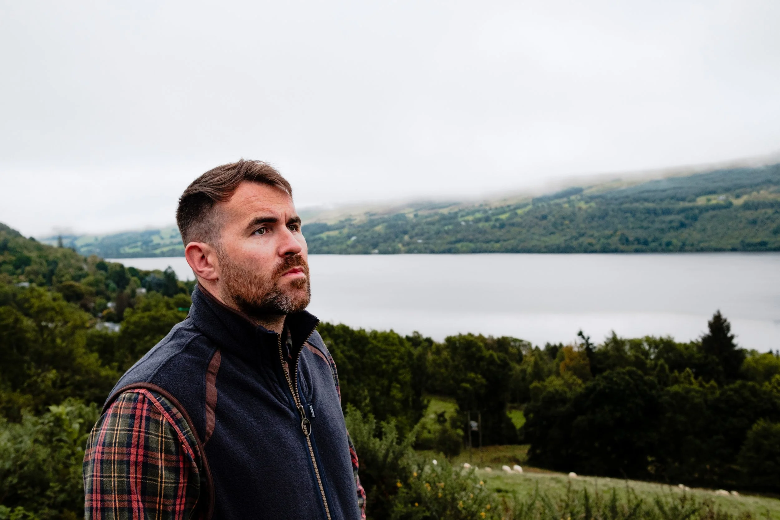 A man with short brown hair and a beard stands outdoors near a lake, with green hills and cloudy sky in the background.