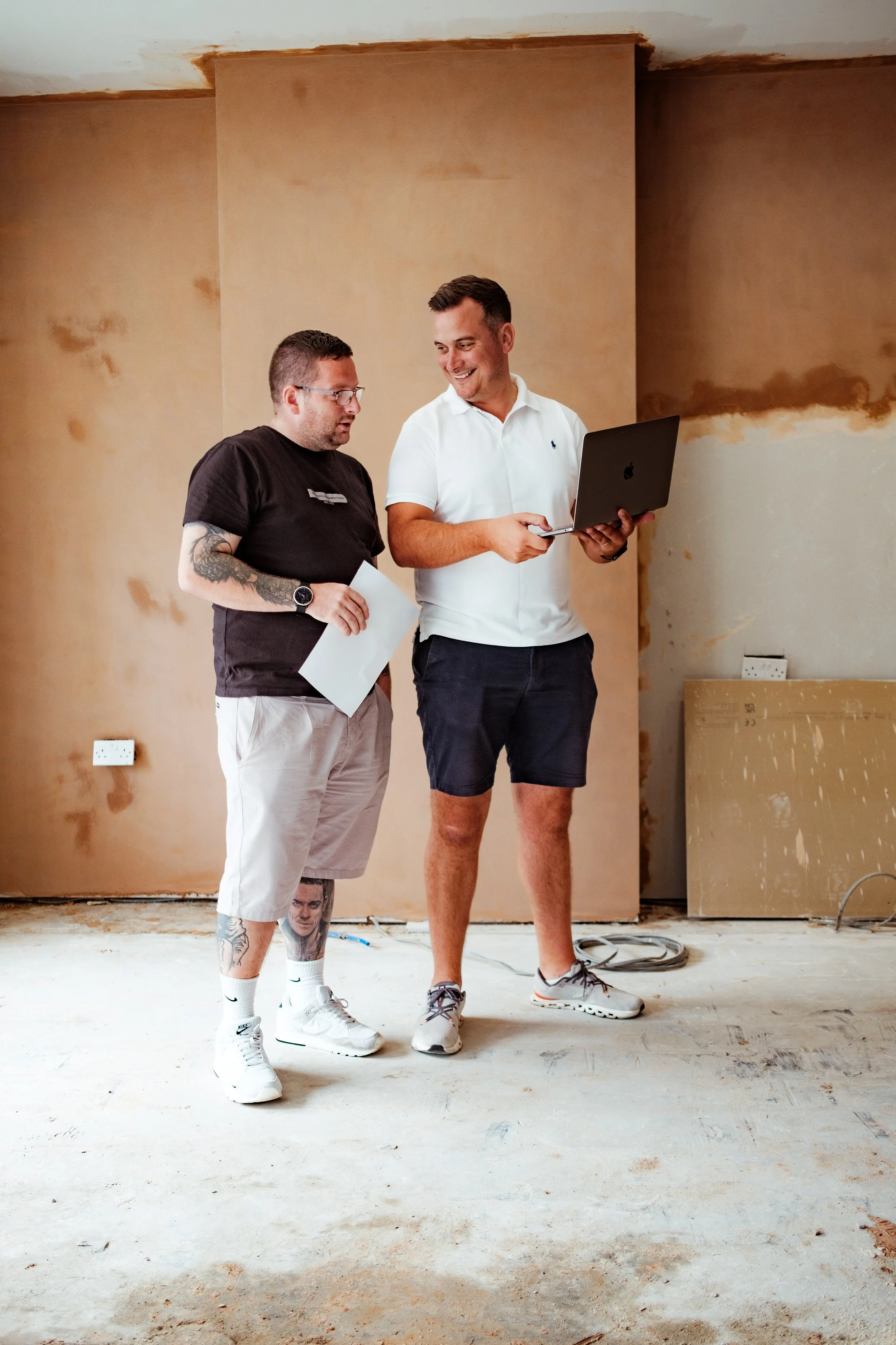 Two men are standing and talking in a room under construction, with unfinished floors and walls, one holding a laptop and the other with a paper.