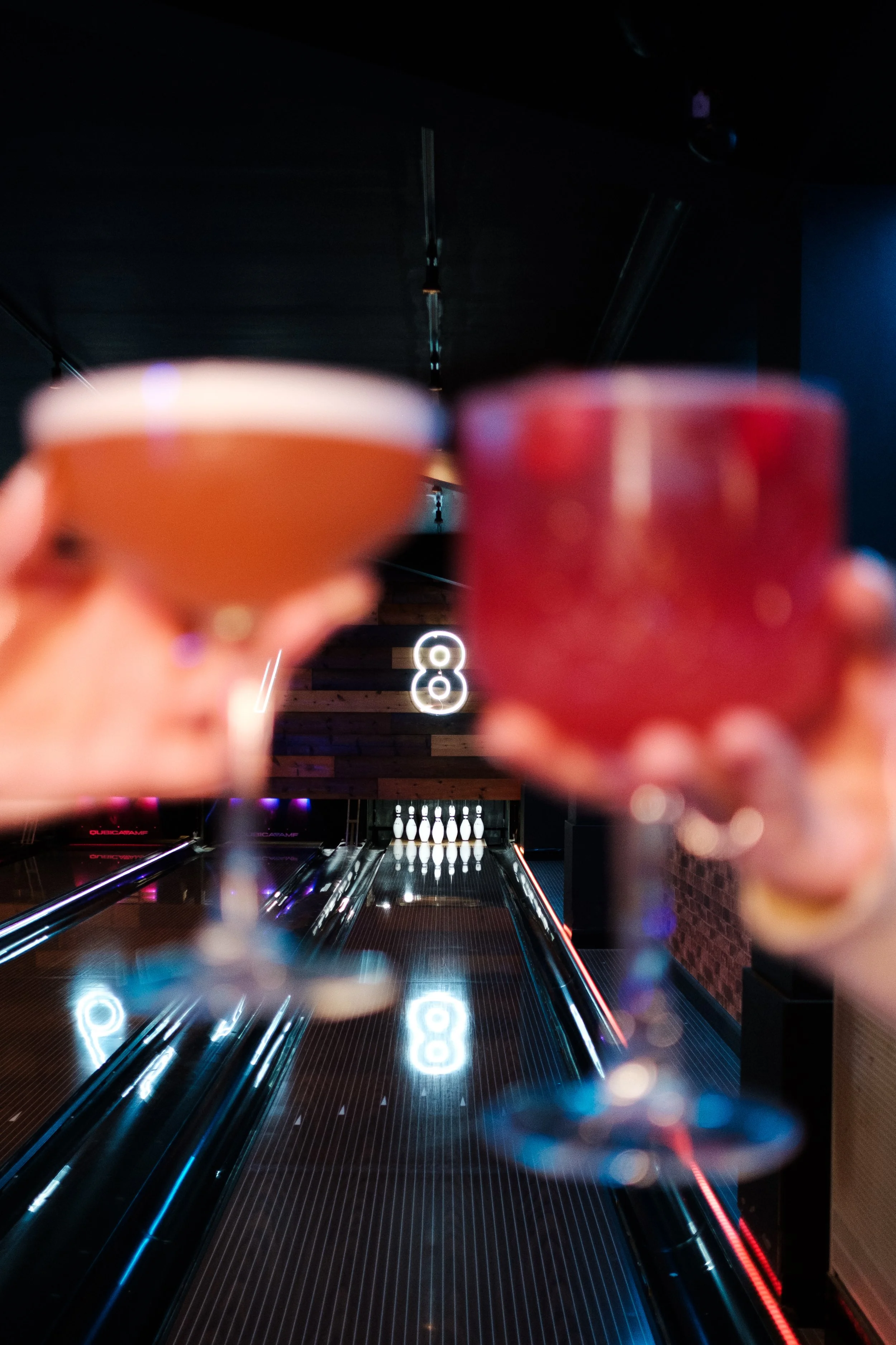 Two cocktails held up in front of a bowling alley with pins and a glowing number 8 in the background.