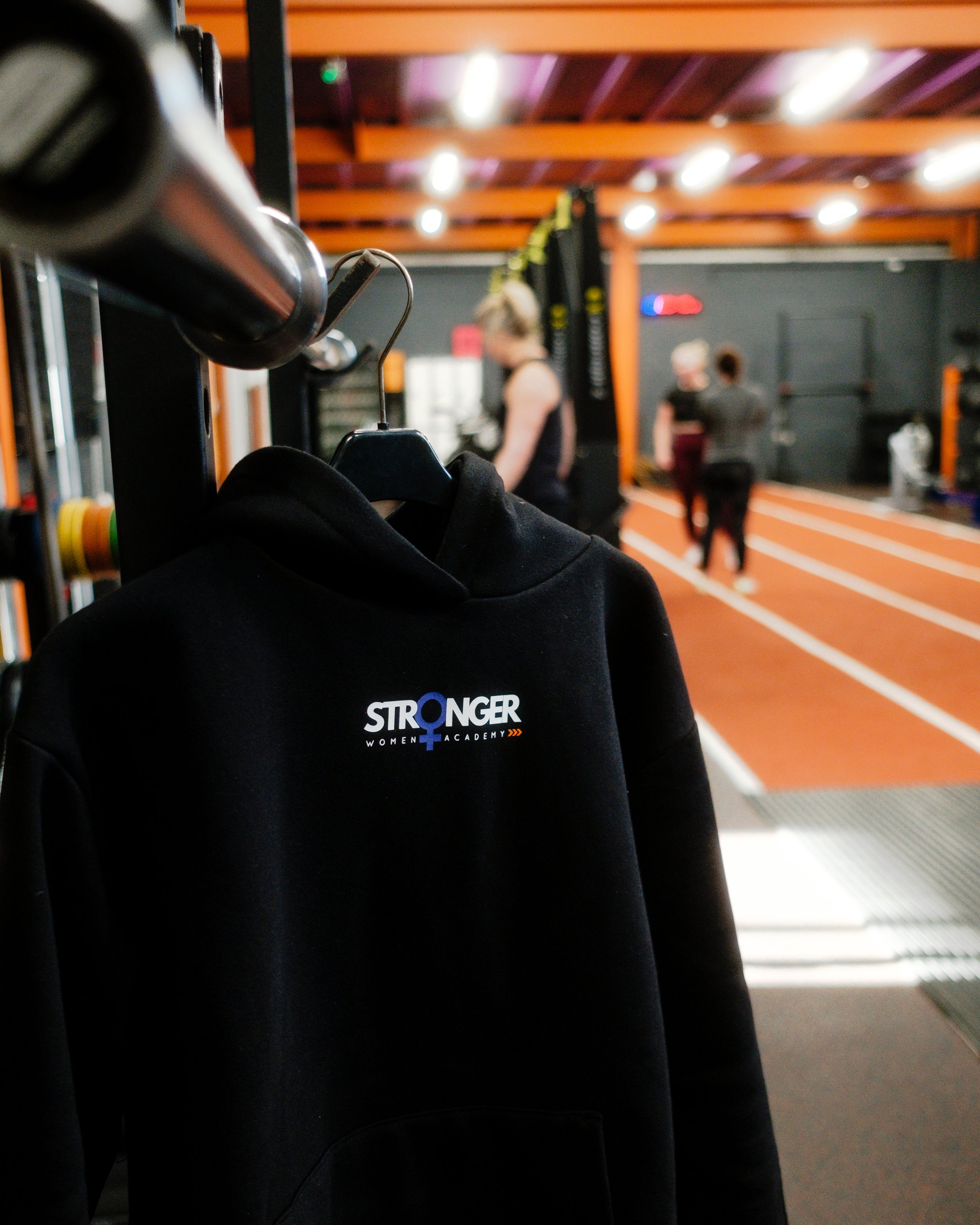 Black hoodie on a hanger with the words 'STRONGER WOMEN'S ACADEMY' printed on it, hanging in a gym with people working out in the background.