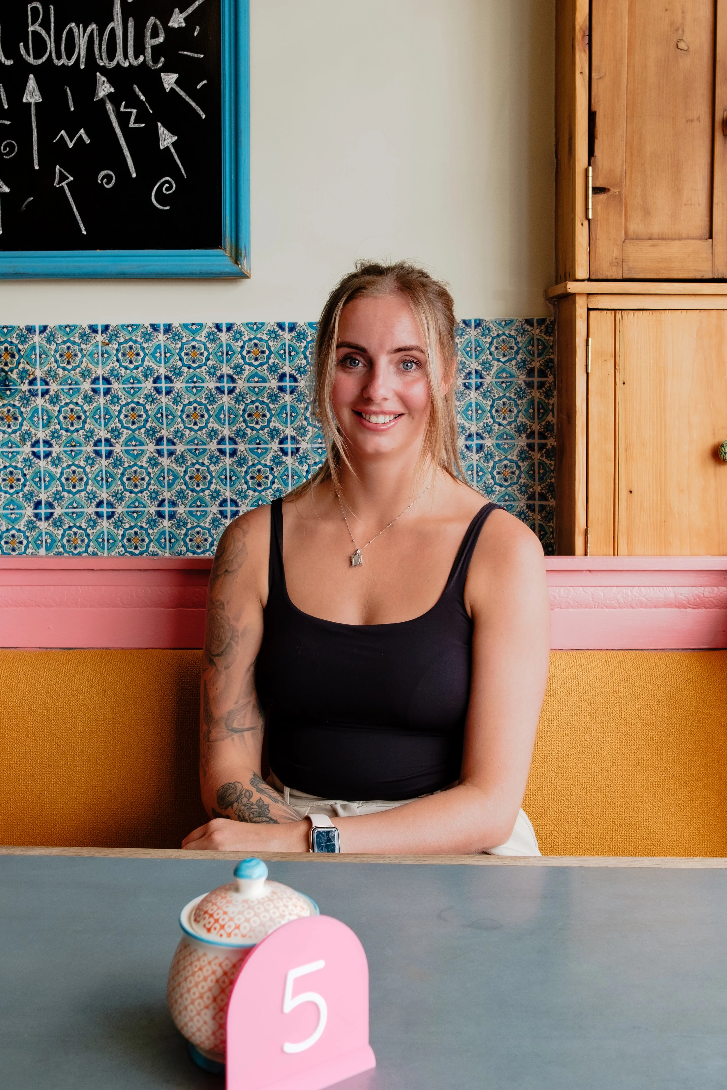 A young woman with blonde hair, tattoos on her right arm, wearing a black tank top, sitting at a table in a colorful cafe, smiling at the camera. In the foreground, there are a decorative container with a lid and a pink table number sign showing the 