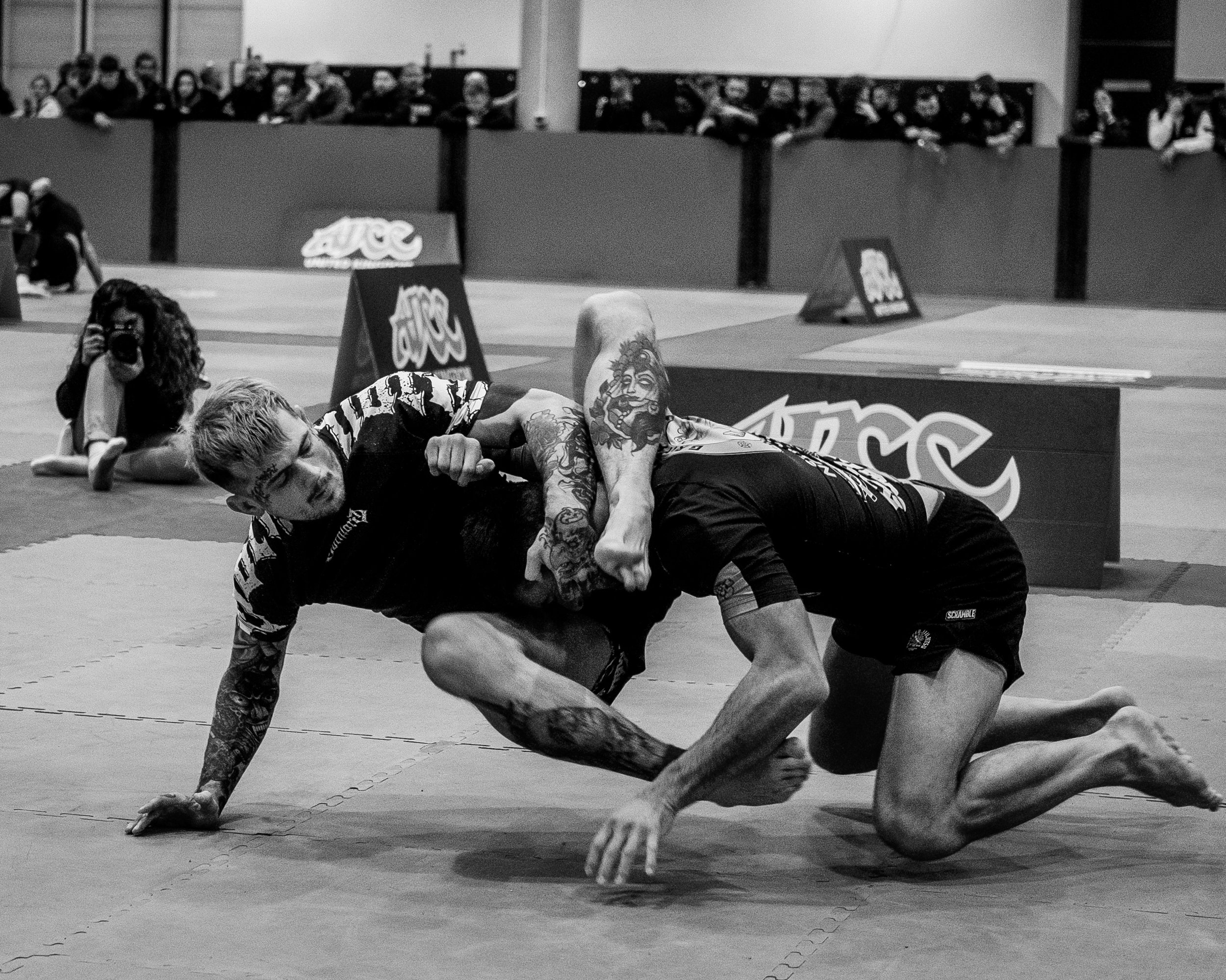Two mixed martial artists competing in a grappling match on a mats, with a photographer capturing the action in the background, and spectators watching from the stands.