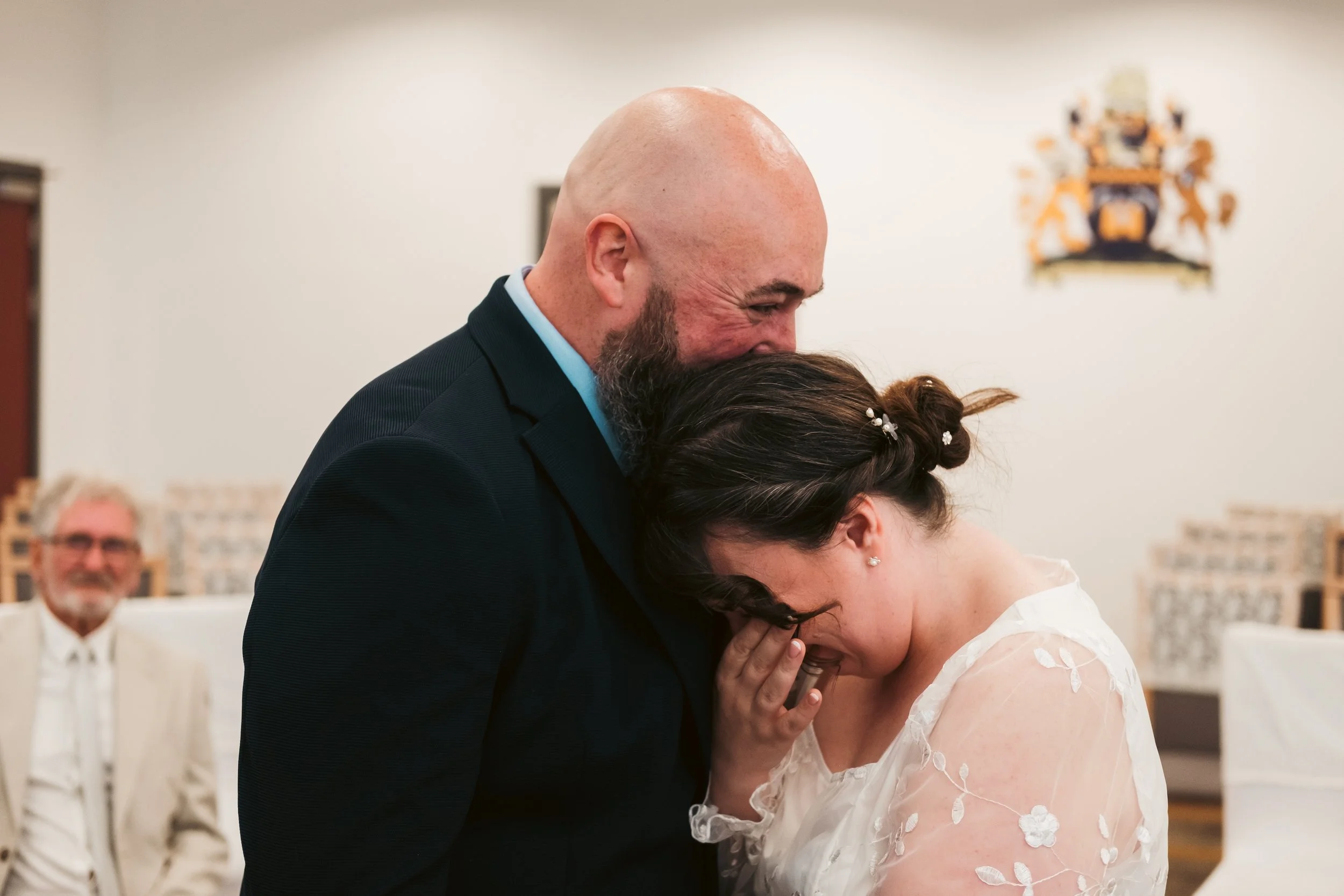 A bald man with a beard and a woman with dark hair in a wedding dress sharing a close, emotional moment during a wedding ceremony.