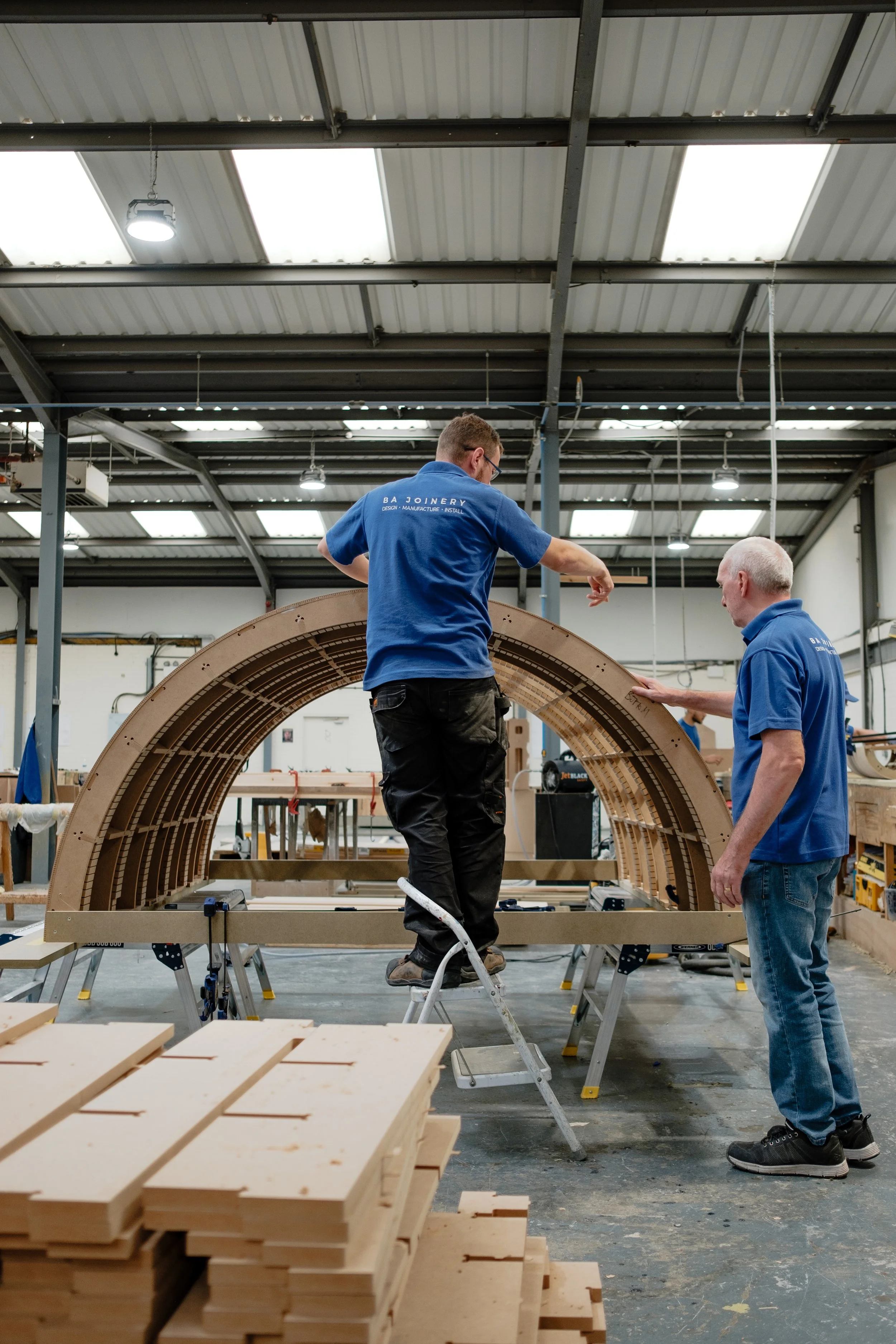 Two workers assembling a large wooden arc in a workshop, with various tools and stacks of wood around them.
