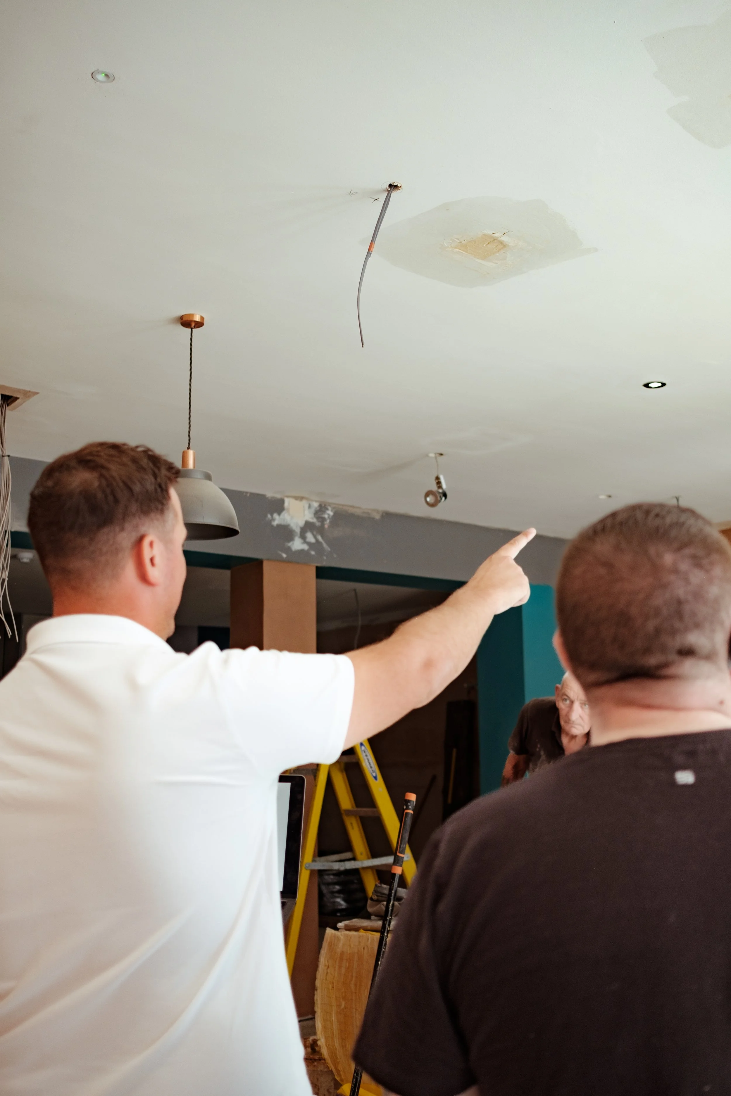 People discussing construction or renovation work indoors, with one person pointing at the ceiling which has visible patches and exposed wiring.