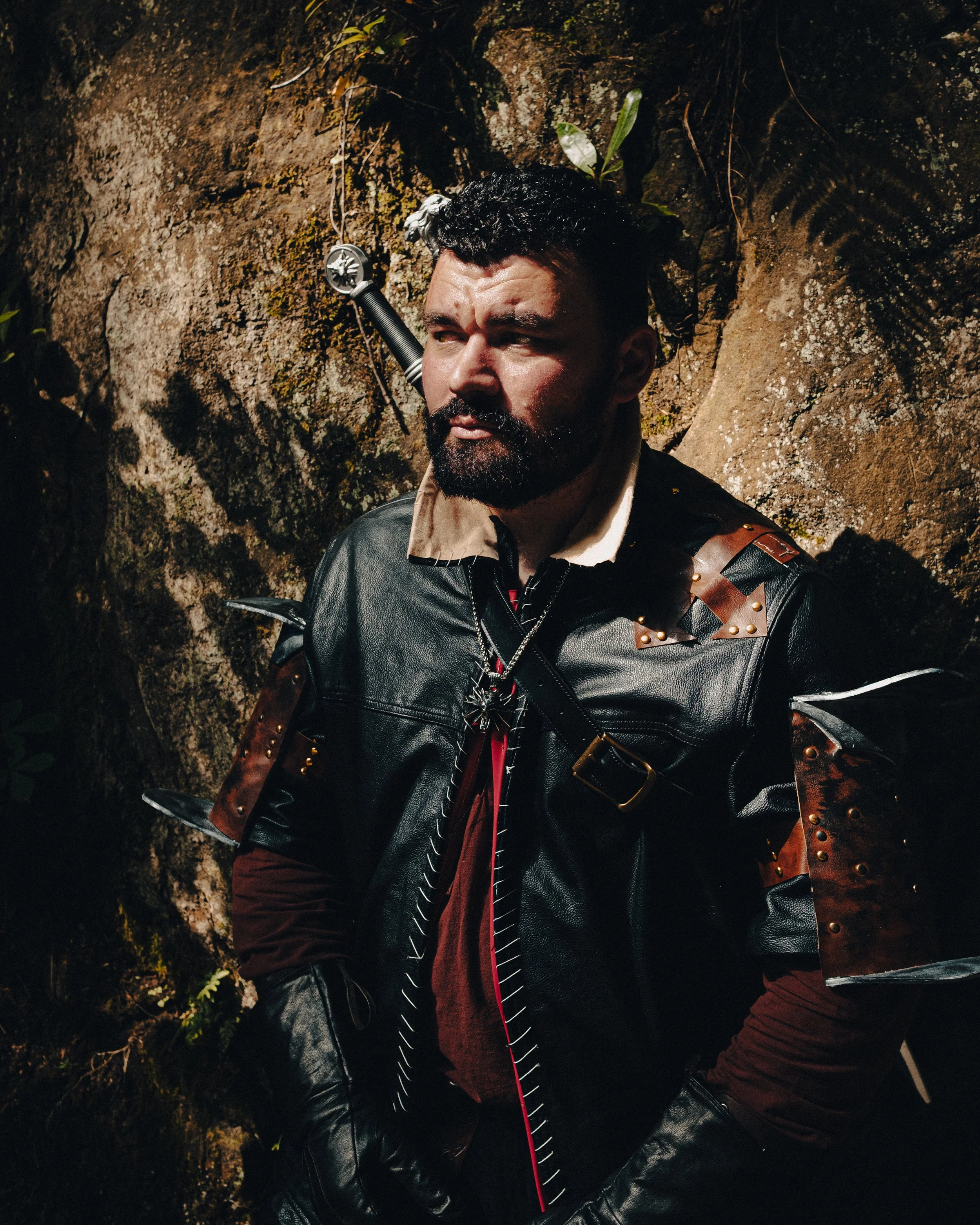 A man with dark curly hair and a beard wearing a black leather jacket with brown leather accents and metal studs, standing outdoors against a rocky background with some green plants.