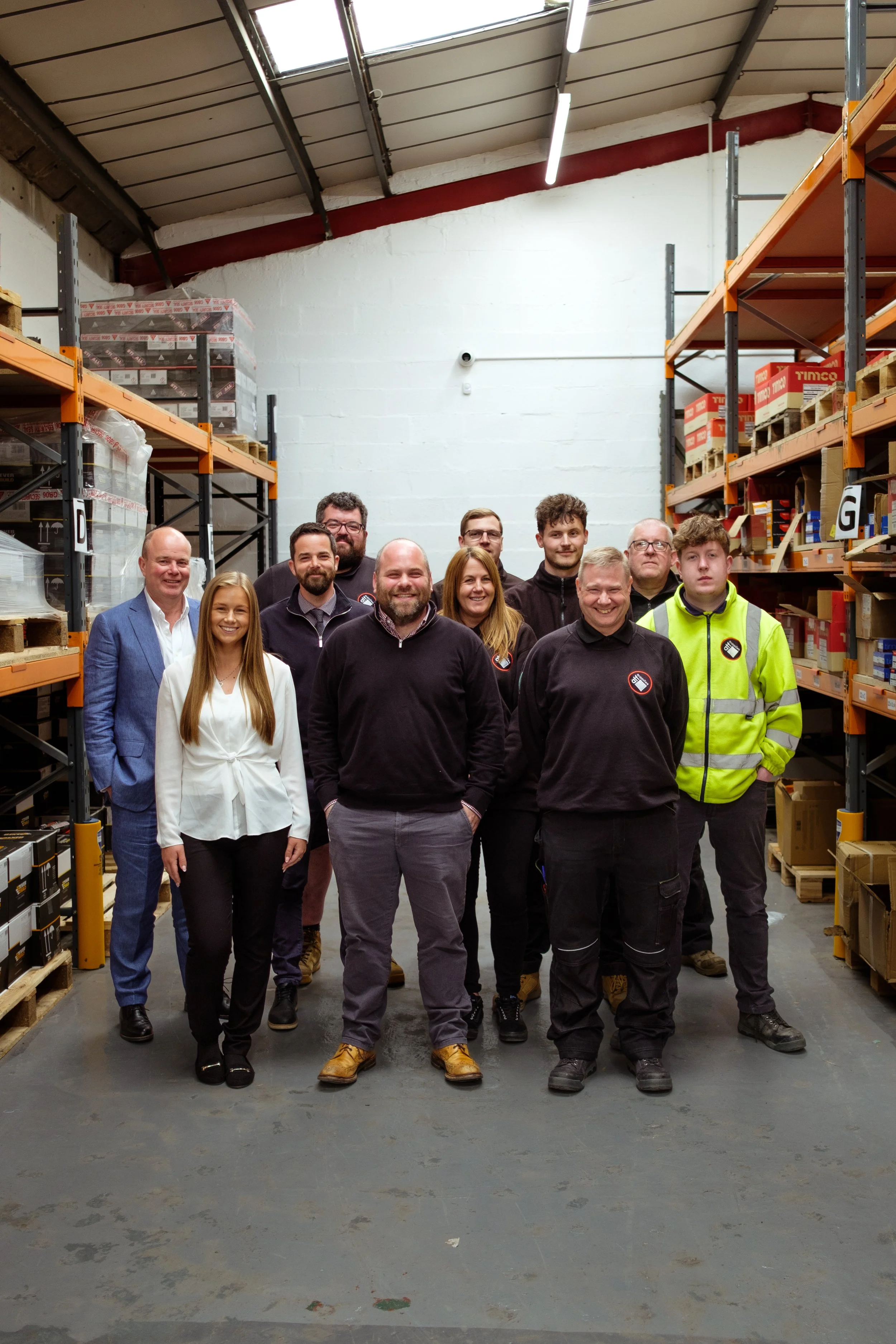 A group of people standing together in a warehouse aisle with shelves of boxes on both sides.