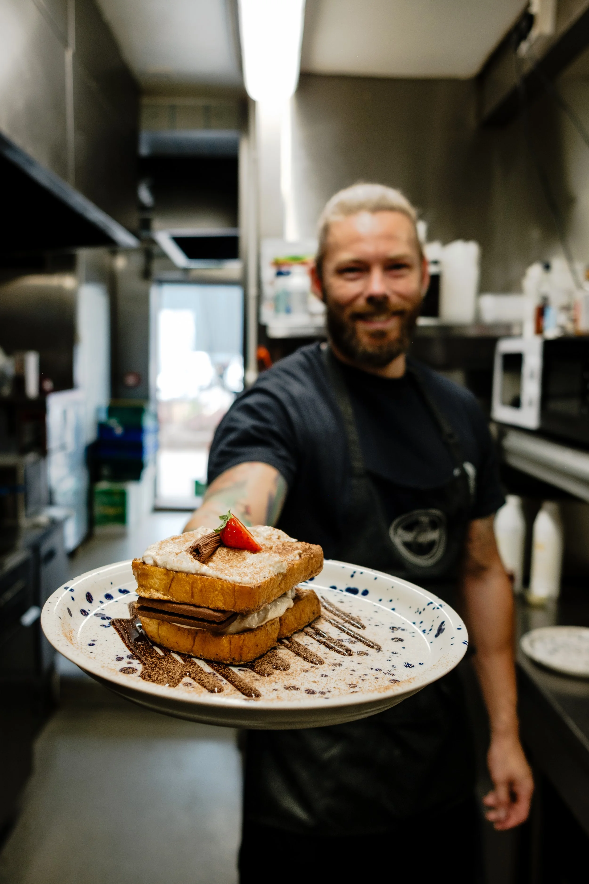 A man holding a plate with three slices of French toast topped with strawberries, chocolate, and powdered sugar in a commercial kitchen.