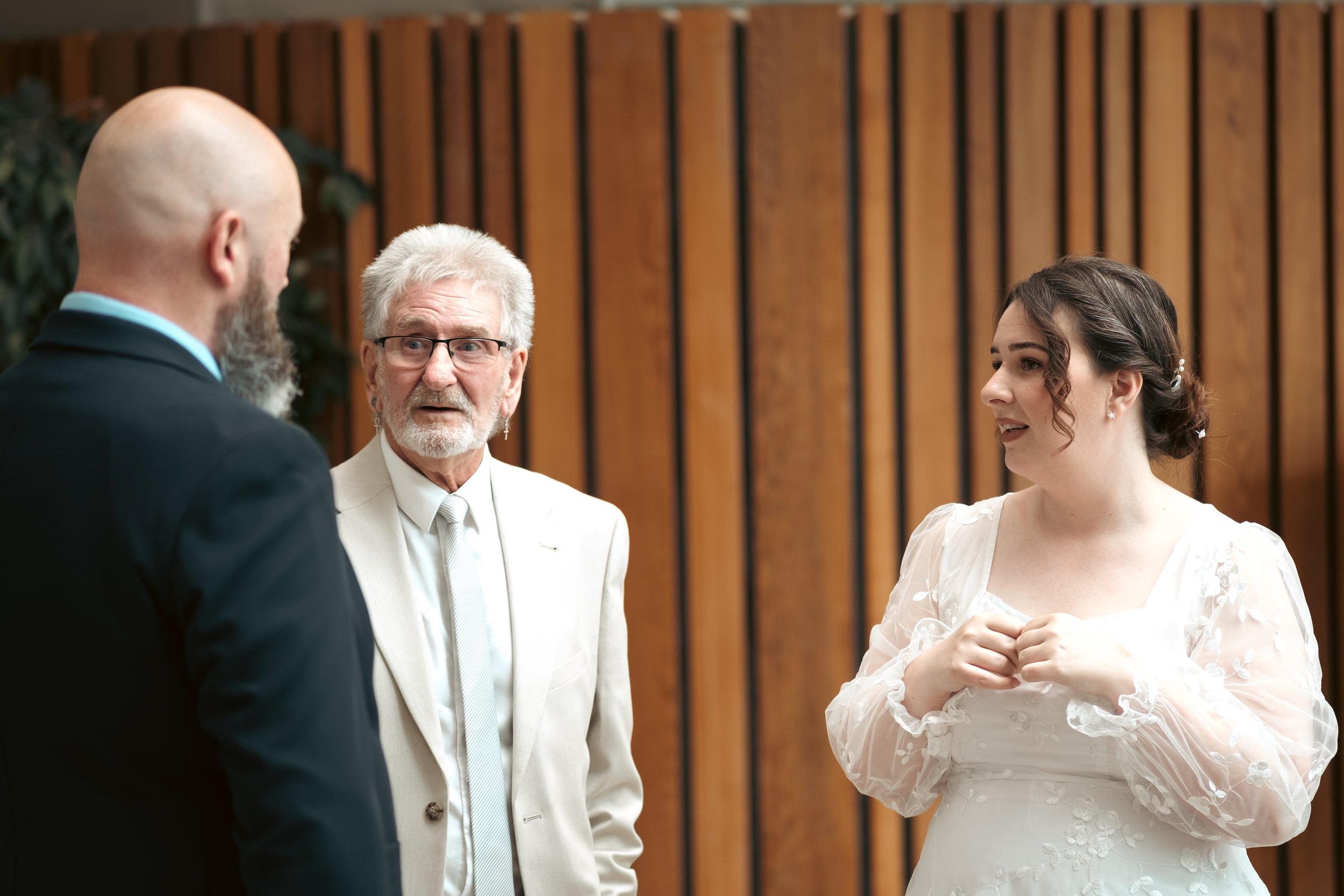 A wedding ceremony with three people, including a person in a white wedding dress, a man in a black suit, and an older man in a white suit with gray hair and glasses, standing in front of a wooden panel wall.