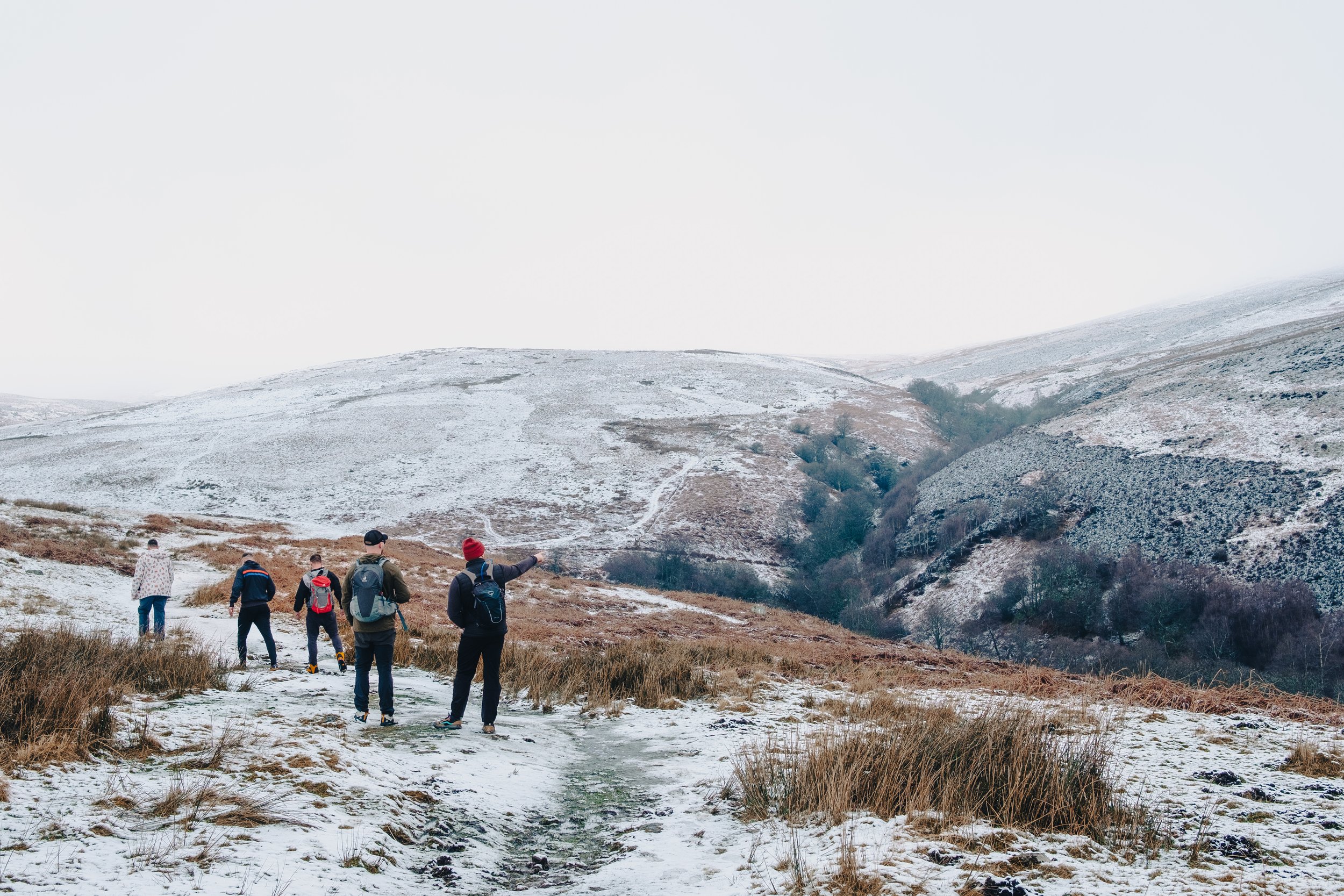A group of six hikers walking on a snowy trail through a hilly landscape with sparse vegetation and snow-covered hills in the background.