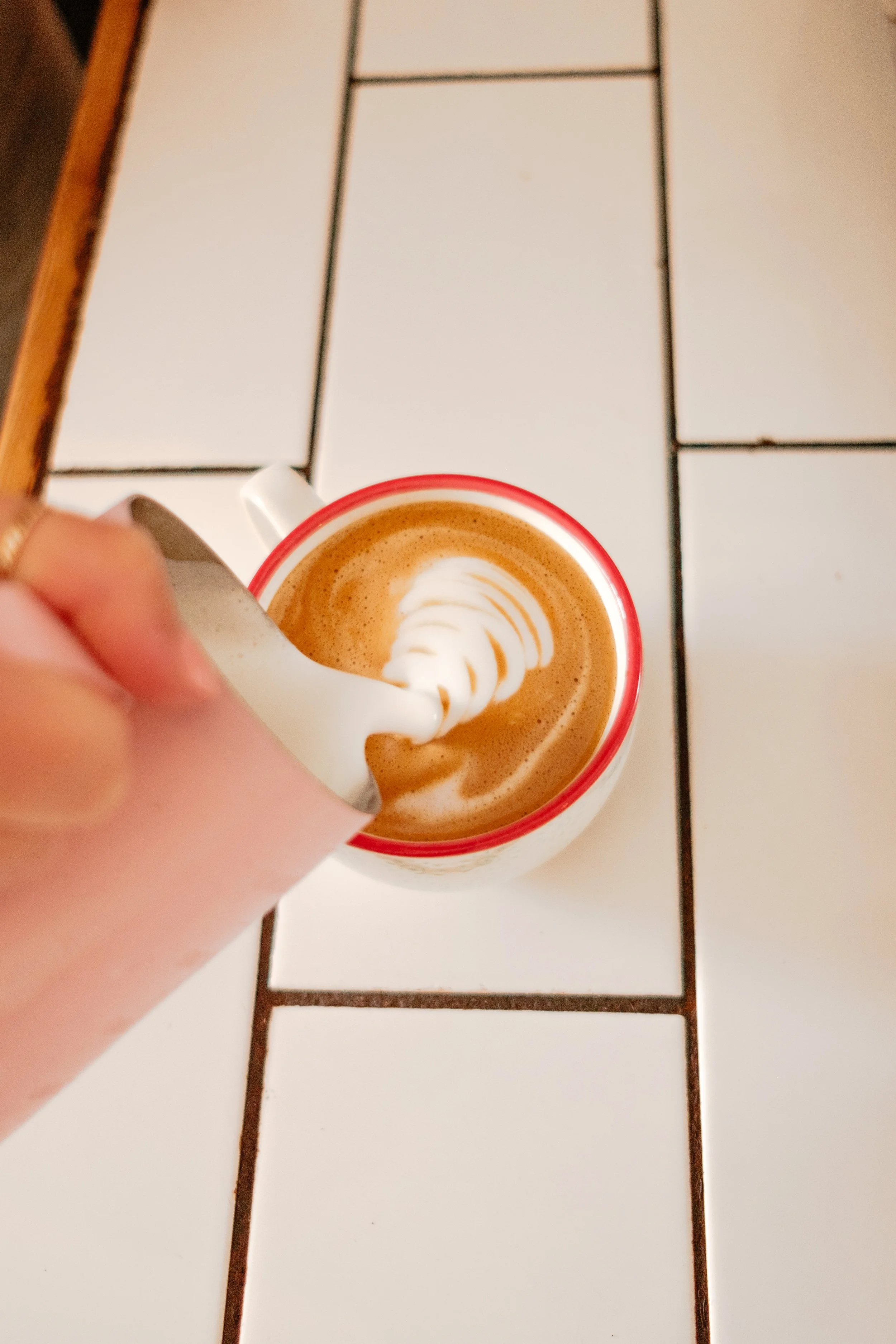 A hand pouring whipped cream into a cup of coffee with latte art, on a tiled table.
