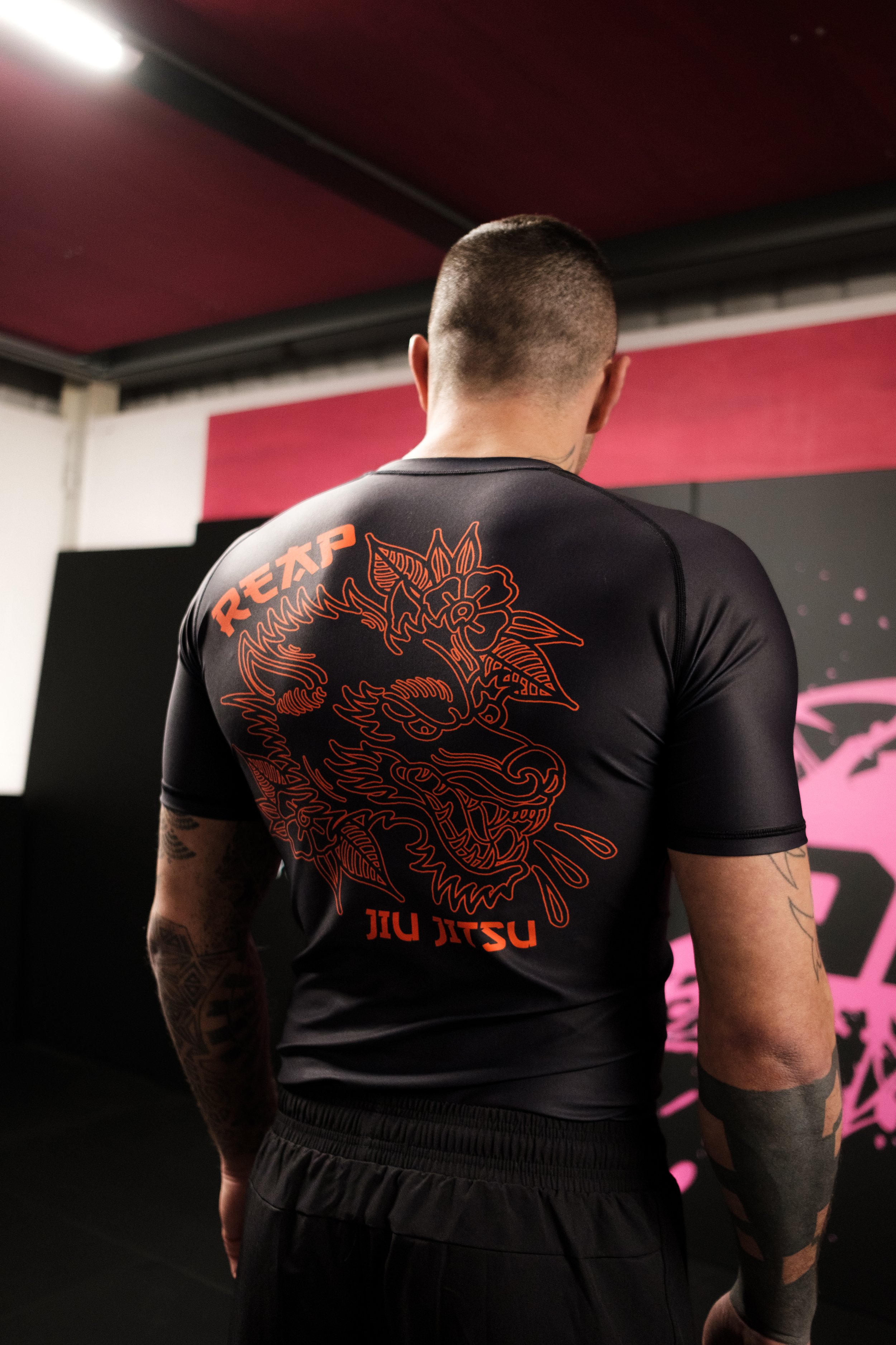 Back view of a person wearing a black rash guard with an orange dragon graphic and text, standing indoors in a martial arts gym.