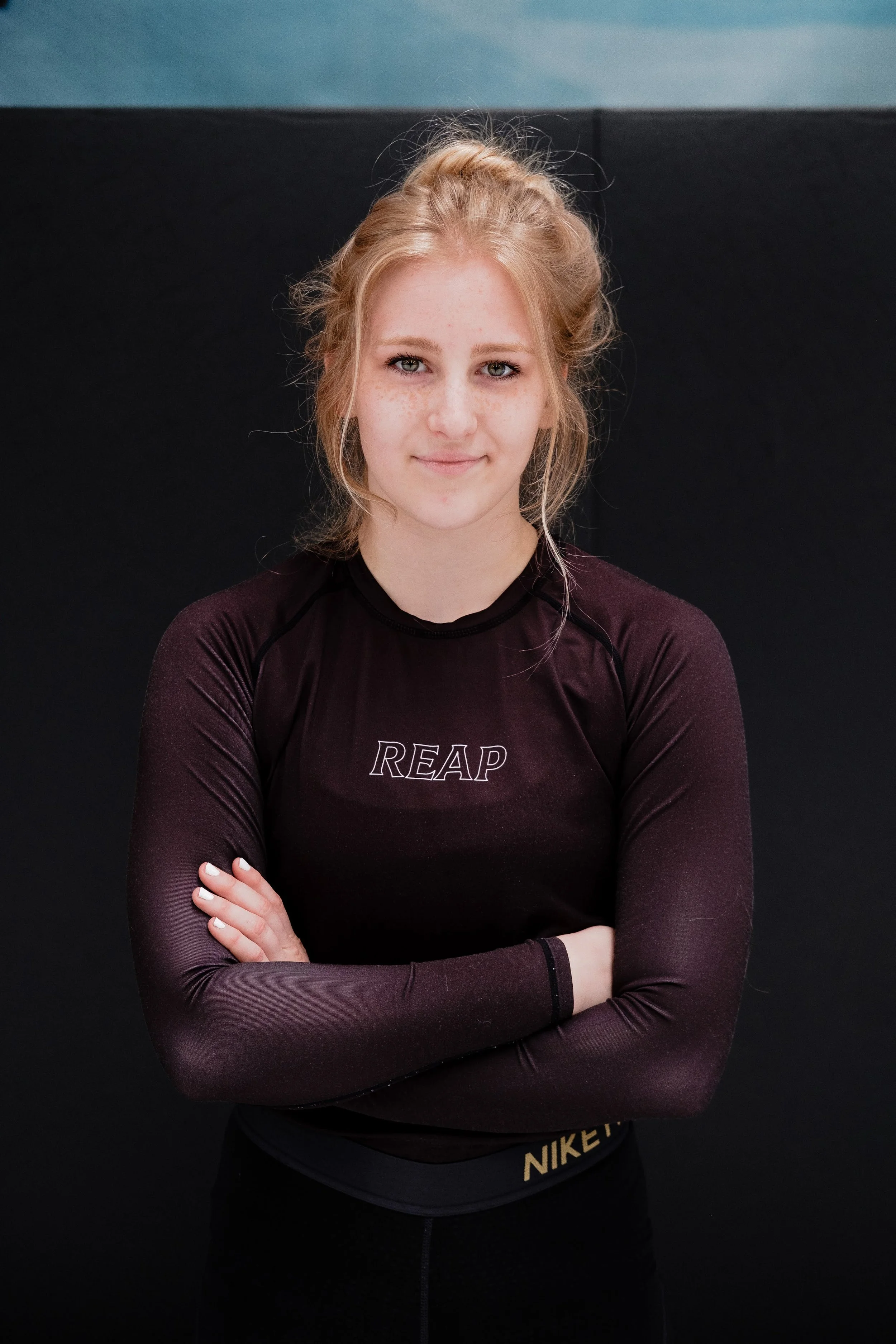 A young woman with light skin, freckles, and blonde hair tied back, standing with her arms crossed, wearing a dark purple athletic top with 'REAP' written on it and black Nike shorts, against a black background.