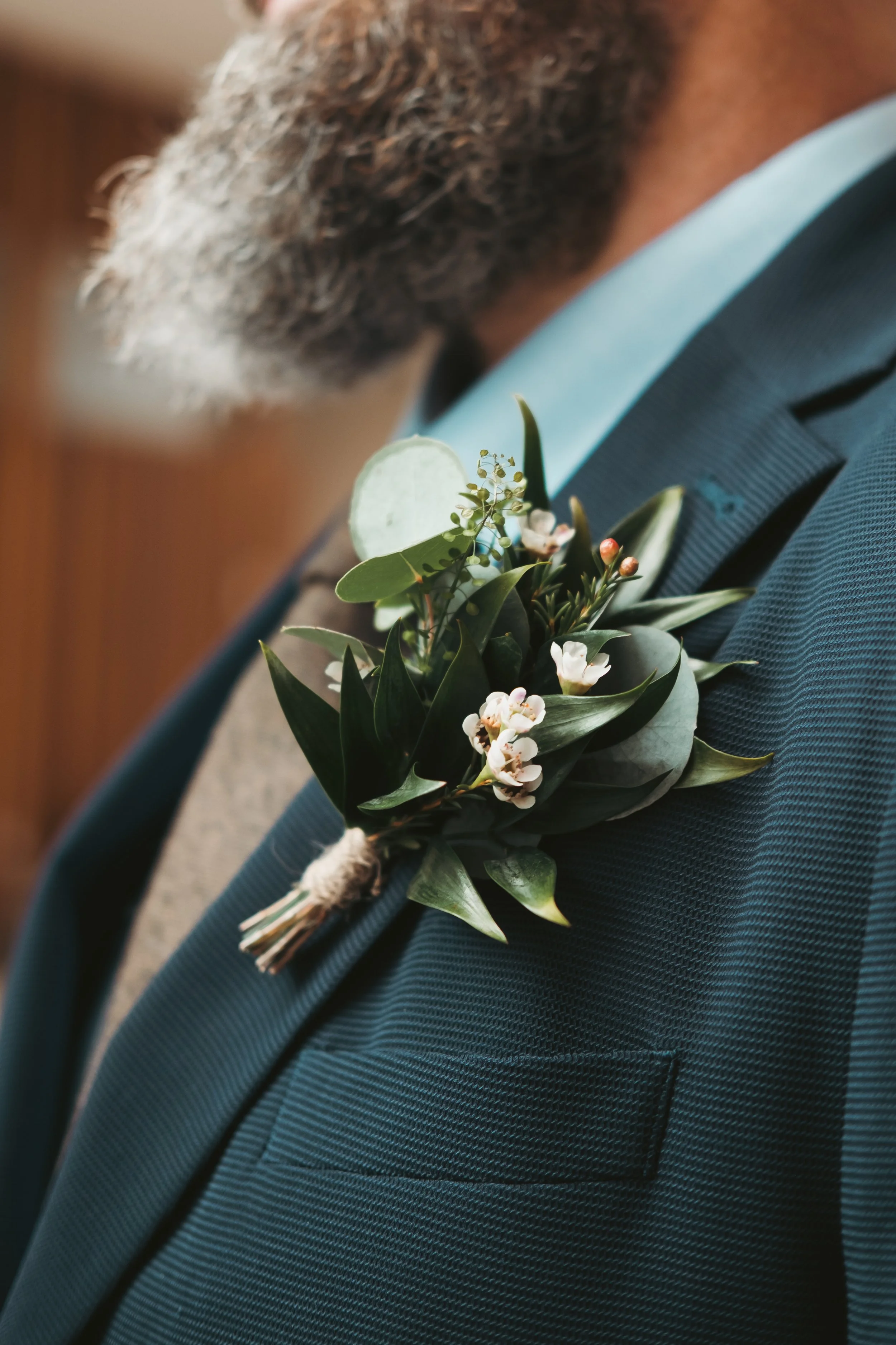 Close-up of a man's suit with a boutonniere of green leaves, small white flowers, and tiny red buds pinned to his lapel. The man has gray curly hair and a beard.
