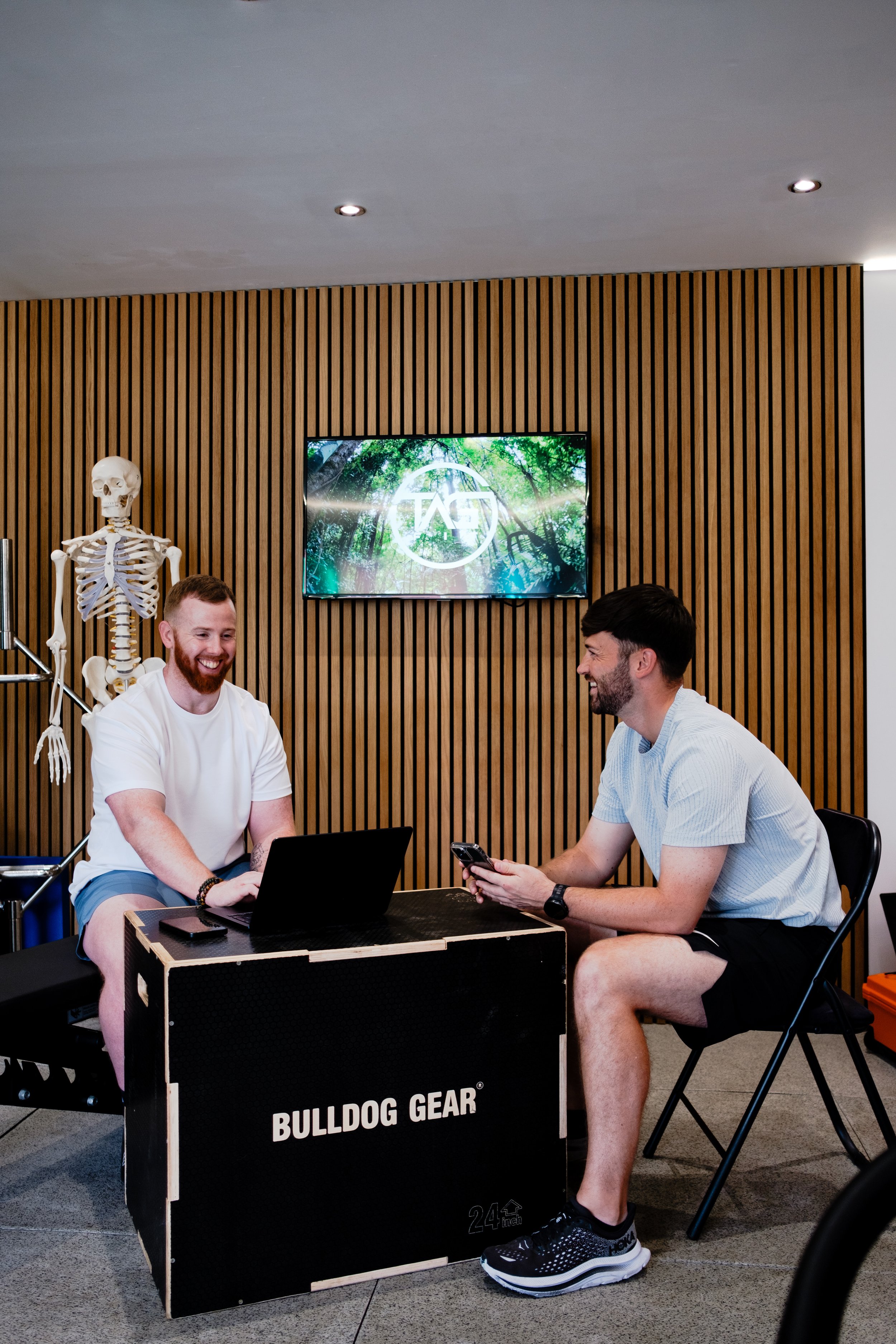 Two men sitting at a table with a laptop and a phone, laughing and talking in an office or fitness environment. A human skeleton model is visible on the left, and a screen is mounted on a wooden slat wall in the background.