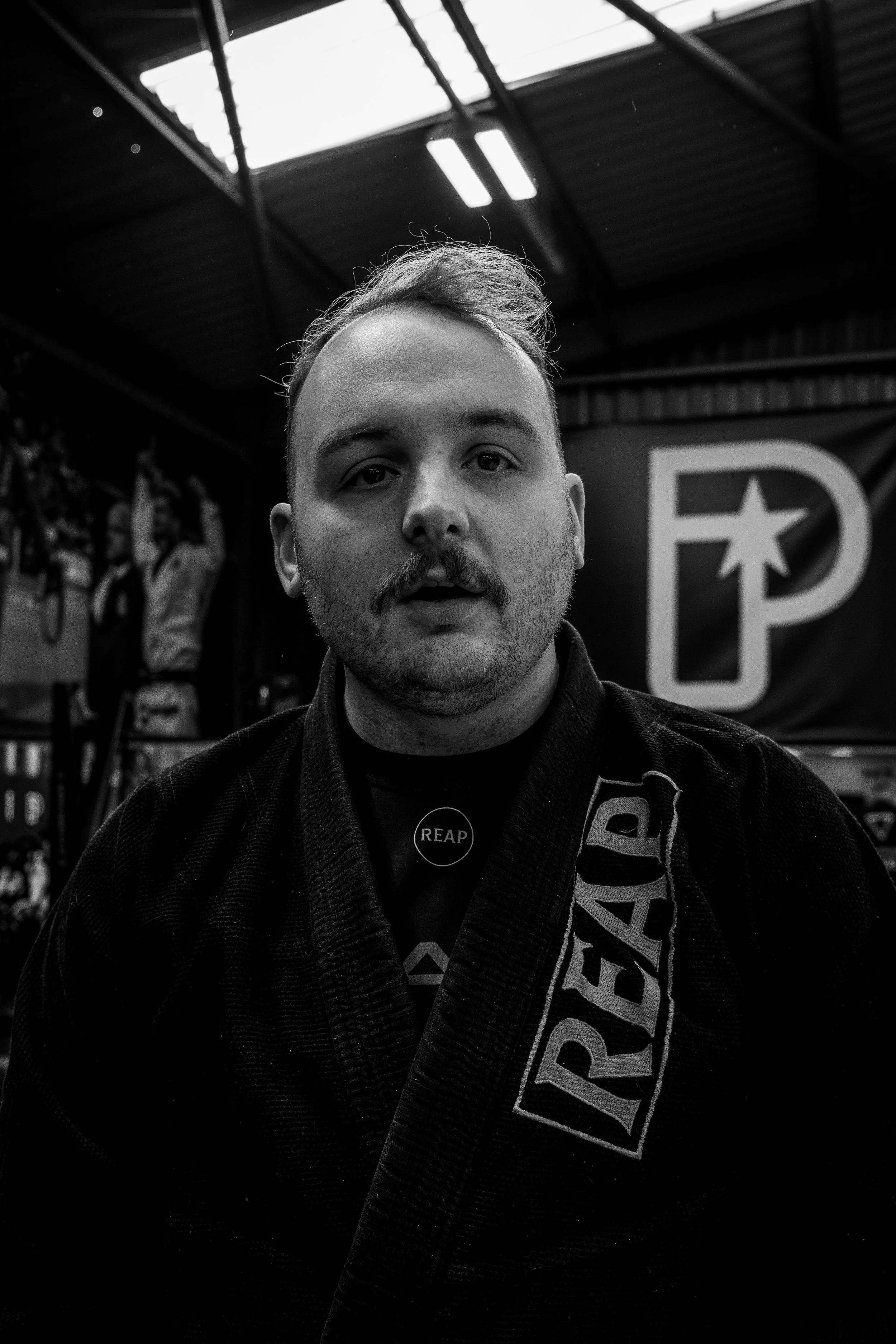 Black and white photo of a man wearing a judo gi with a belt that has the word 'PRIDE' on it, inside a gym or training facility. The background shows sports equipment and a banner with a star and letter 'P'.