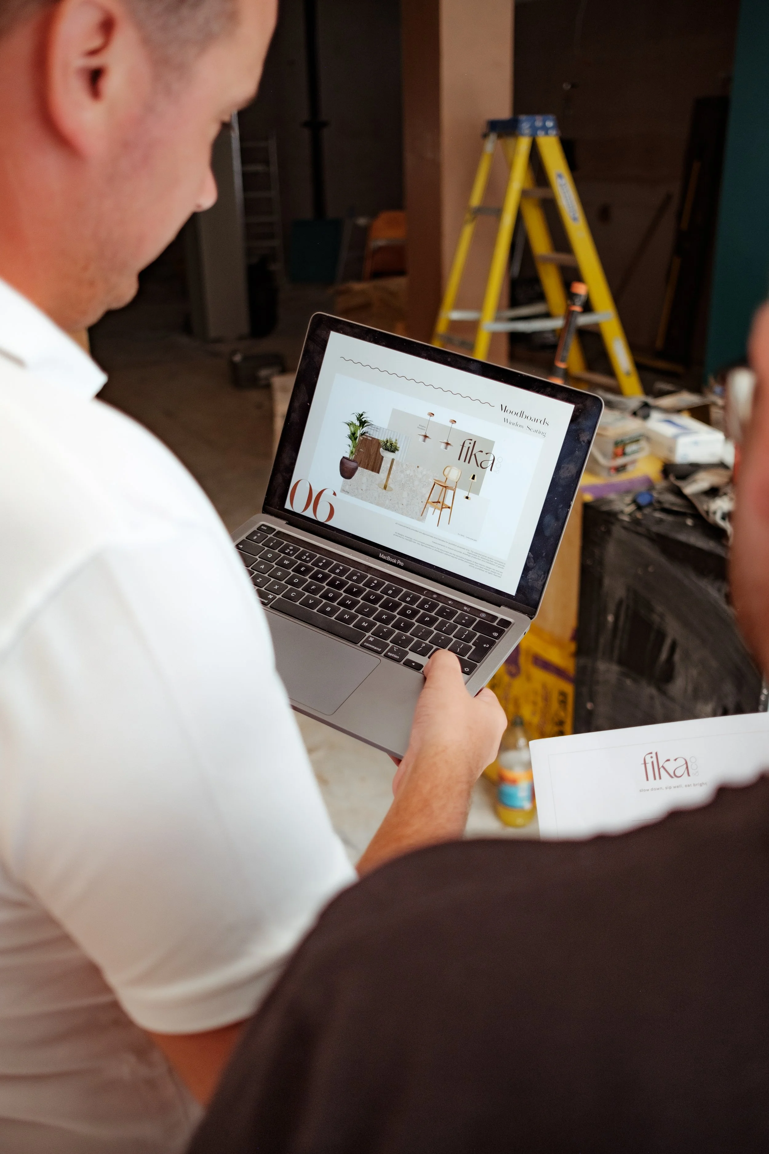 Person holding a laptop showing a design catalog with the title 'Fika' in a workspace with a ladder and tools in the background.