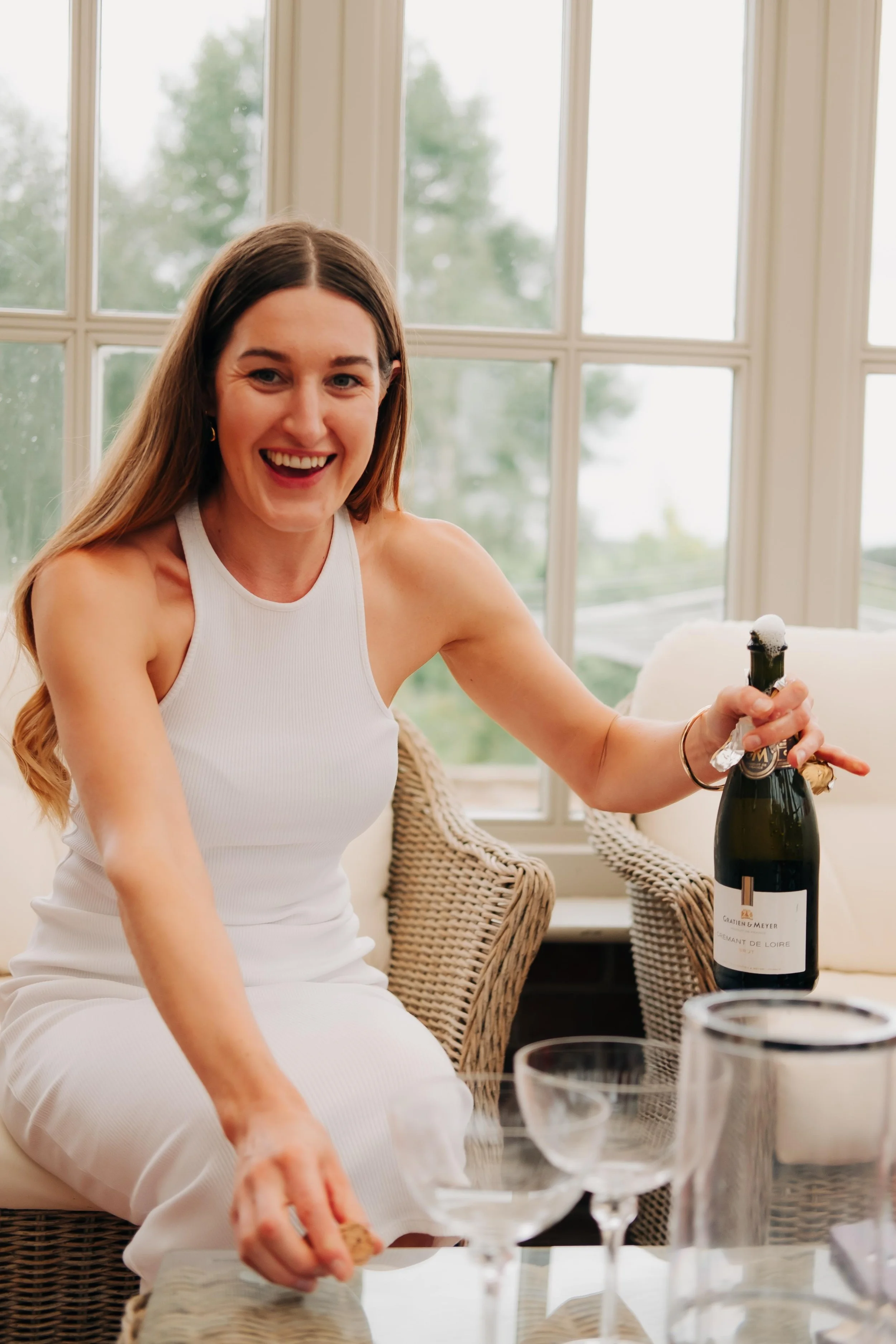 A woman with long brown hair, wearing a white sleeveless dress, smiling and sitting in a wicker chair near a window. She is holding a bottle of sparkling wine or champagne, and there are wine glasses on the table in front of her. The setting appears 