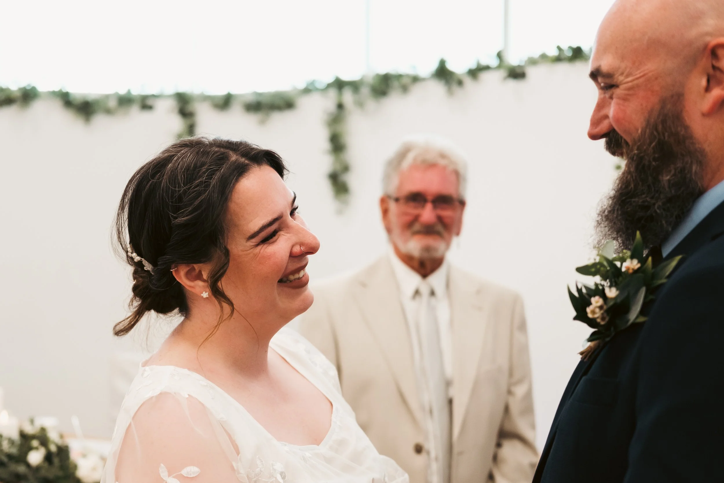 A bride and groom smiling at each other during their wedding ceremony, with an officiant in the background.