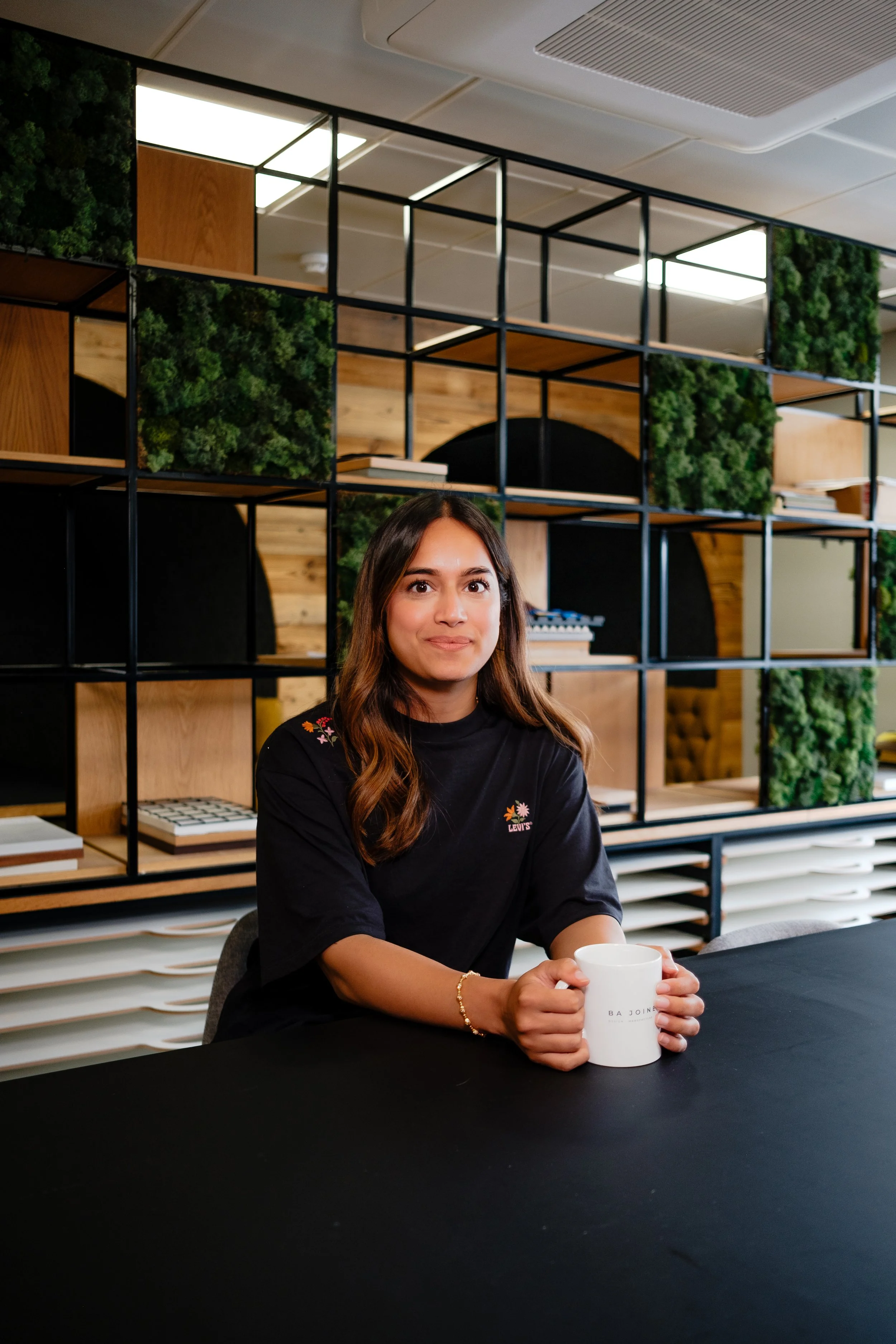 A young woman with long brown hair sitting at a black table in an office, holding a white mug, with modern shelving and green plants in the background.