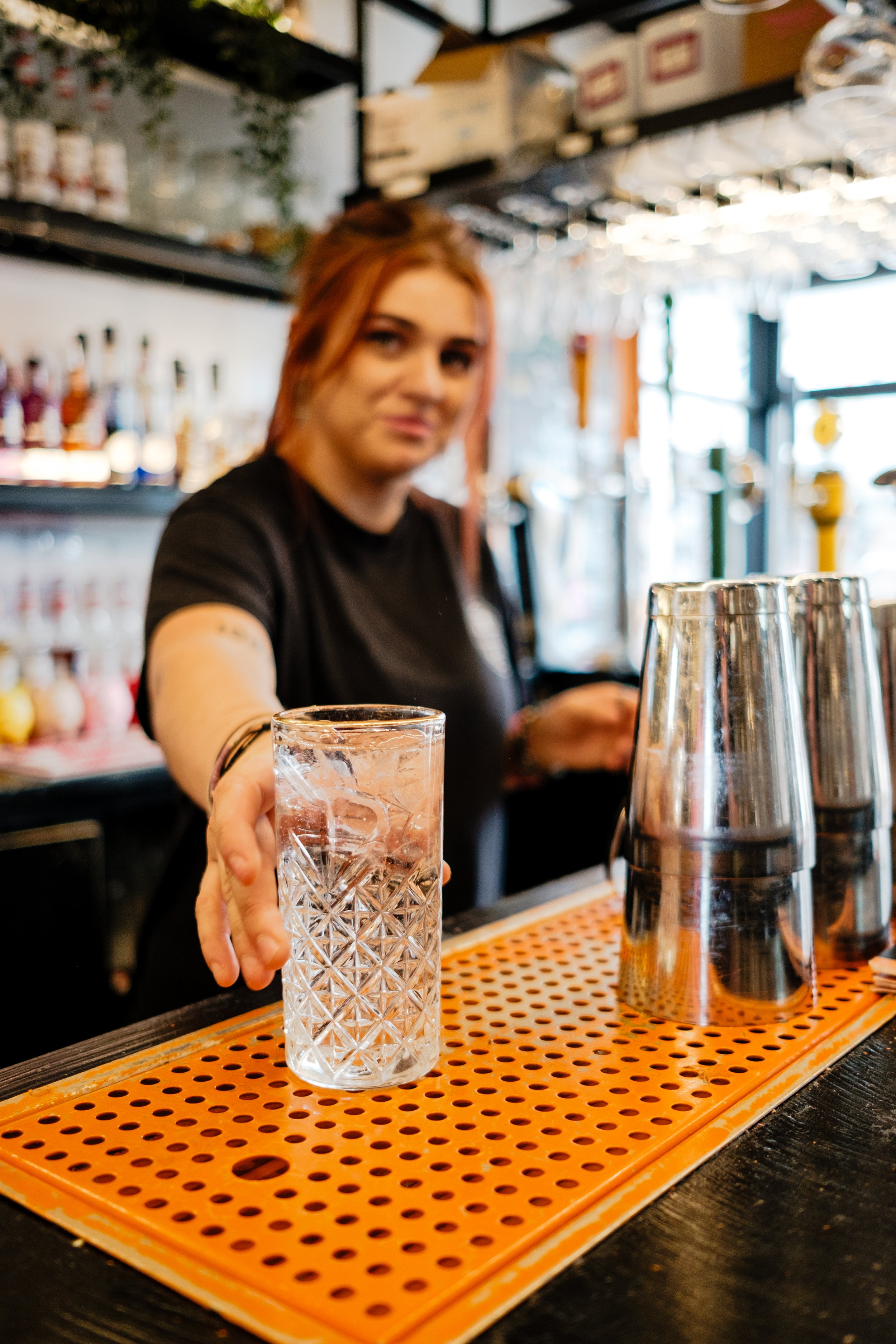 A bartender with reddish hair extending a glass of ice water across the bar counter.