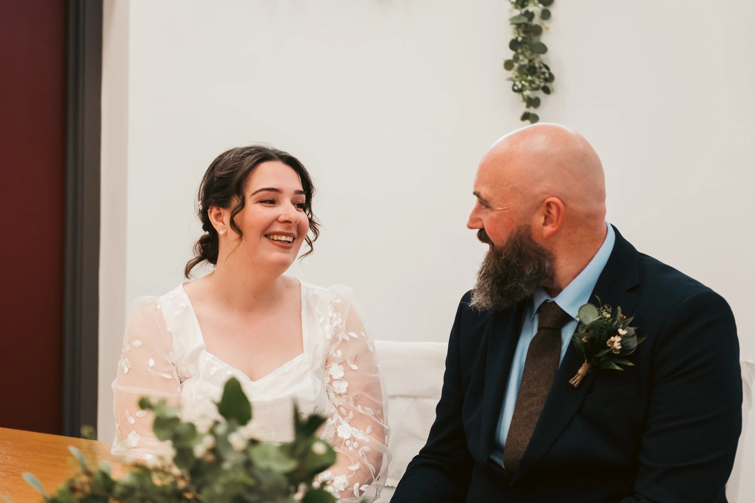 A bride and groom smiling and looking at each other during their wedding ceremony, sitting indoors with white walls and a wall hanging of green leaves in the background.