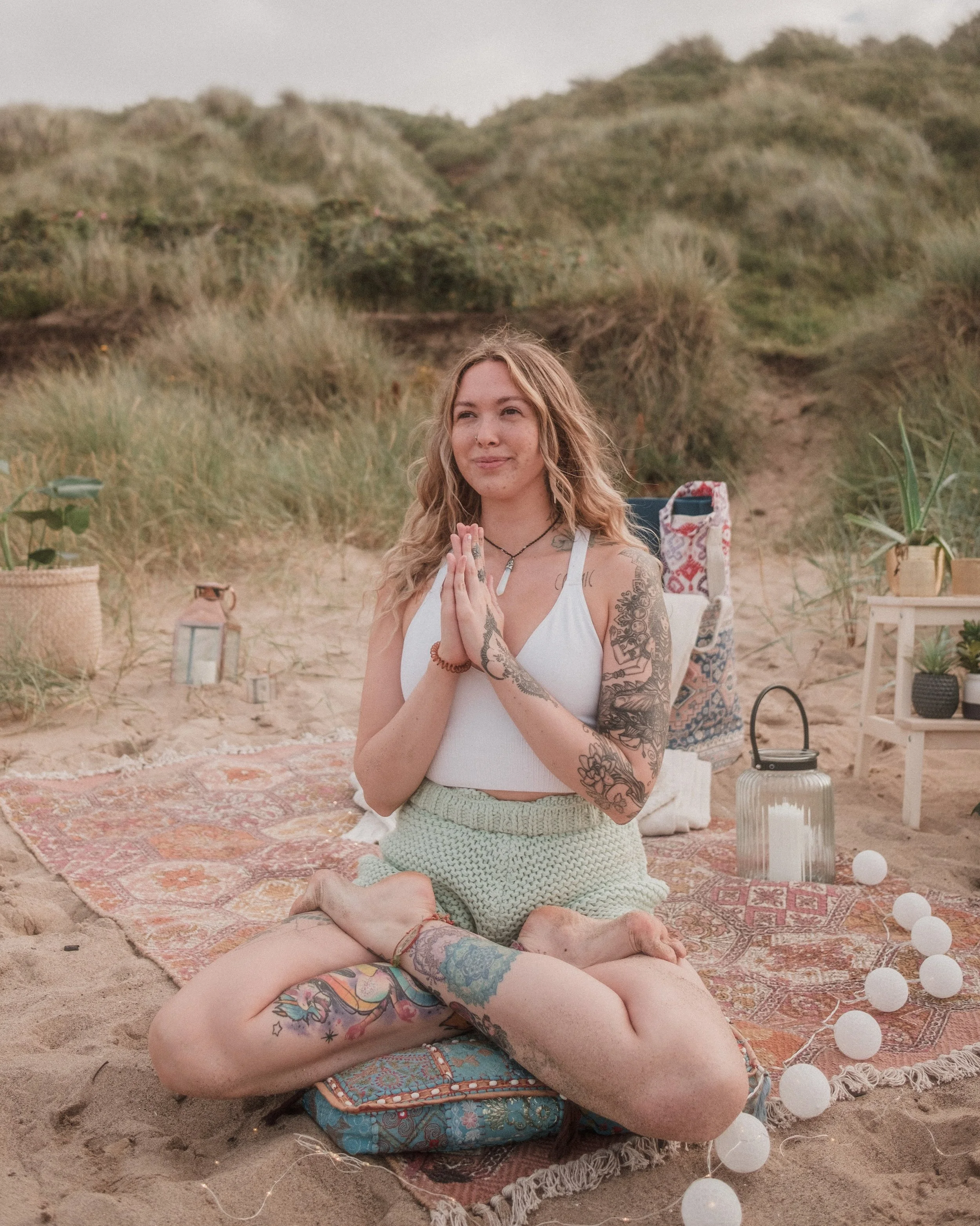 A woman with tattoos on her arms and leg, sitting on the ground in a kneeling position with her hands in a prayer pose, practicing yoga or meditation on a patterned rug at the beach with sand, decorative lights, and a background of green hills.