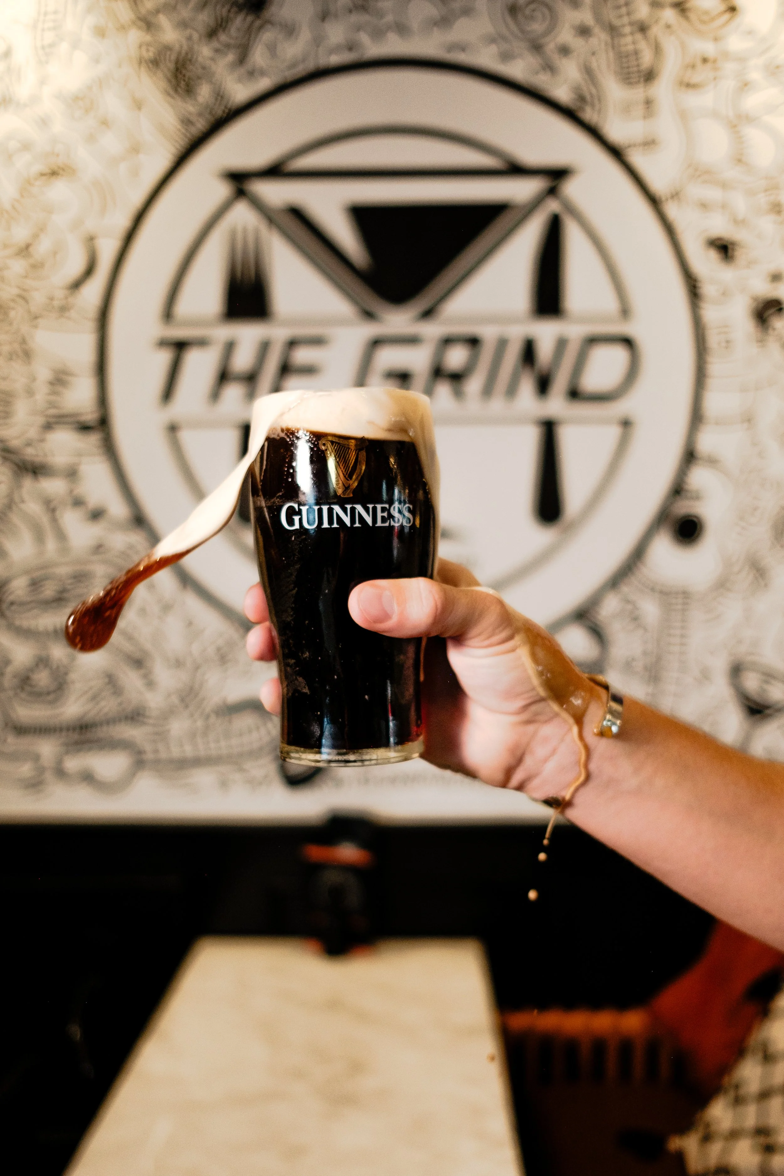 A person holds a Guinness beer glass with dark beer and a thick foamy head in front of a wall with a large, round logo that says 'The Grind'. The person's hand has a ring and a bracelet, and the background appears to be inside a bar or restaurant.