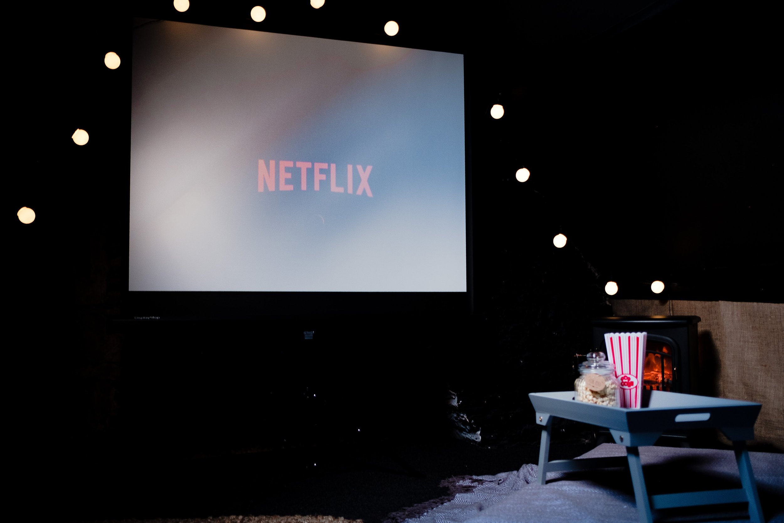 A room with a large screen displaying the Netflix logo, surrounded by string lights. In the foreground, a table with popcorn and a striped popcorn bucket, and a white chair.