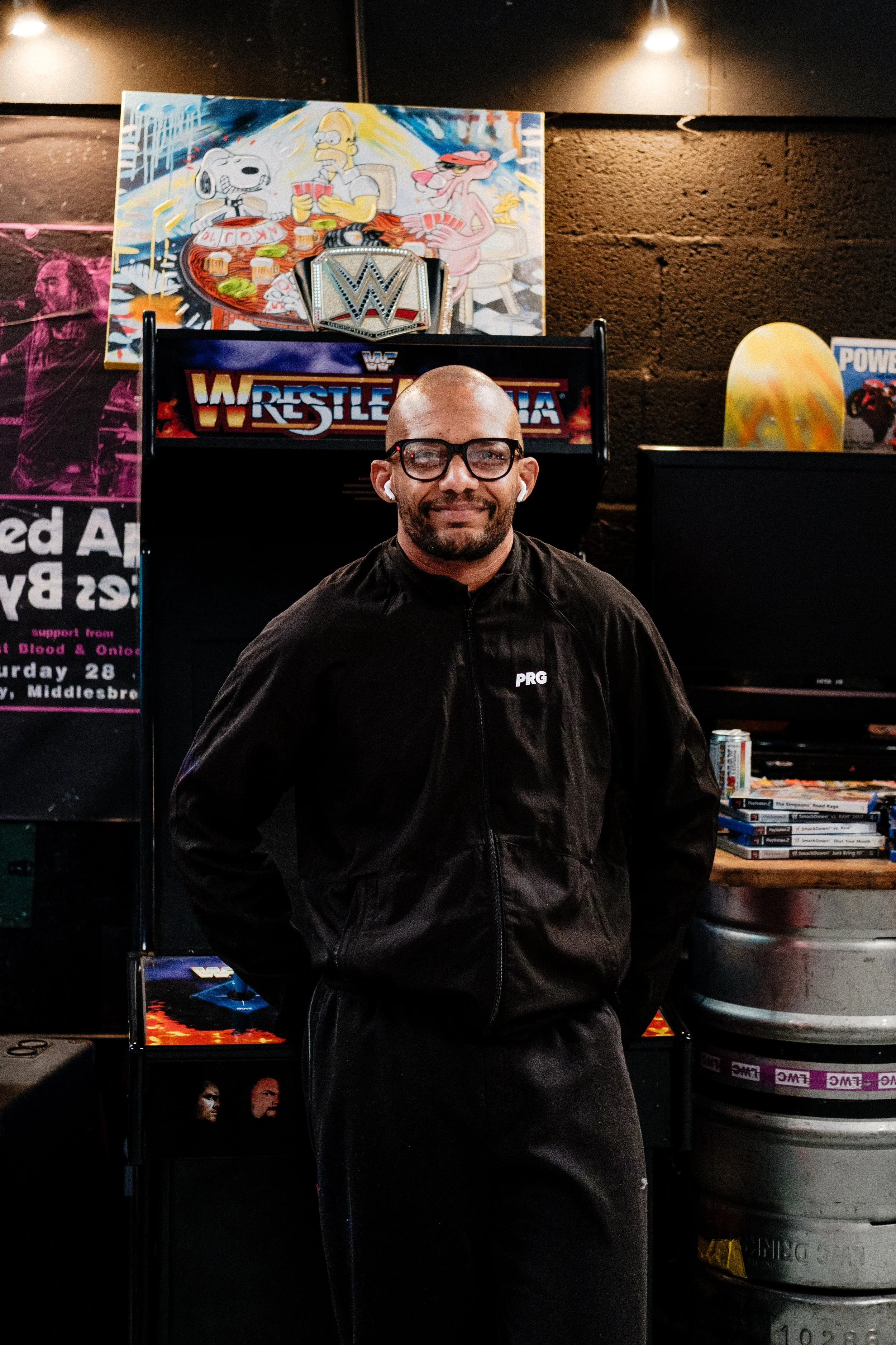 A man with glasses and earbuds standing in front of a WrestleMania arcade game, with a WWE championship belt on top of the game, and a colorful cartoon mural of Homer Simpson, Snoopy, and a pink creature playing poker behind him.