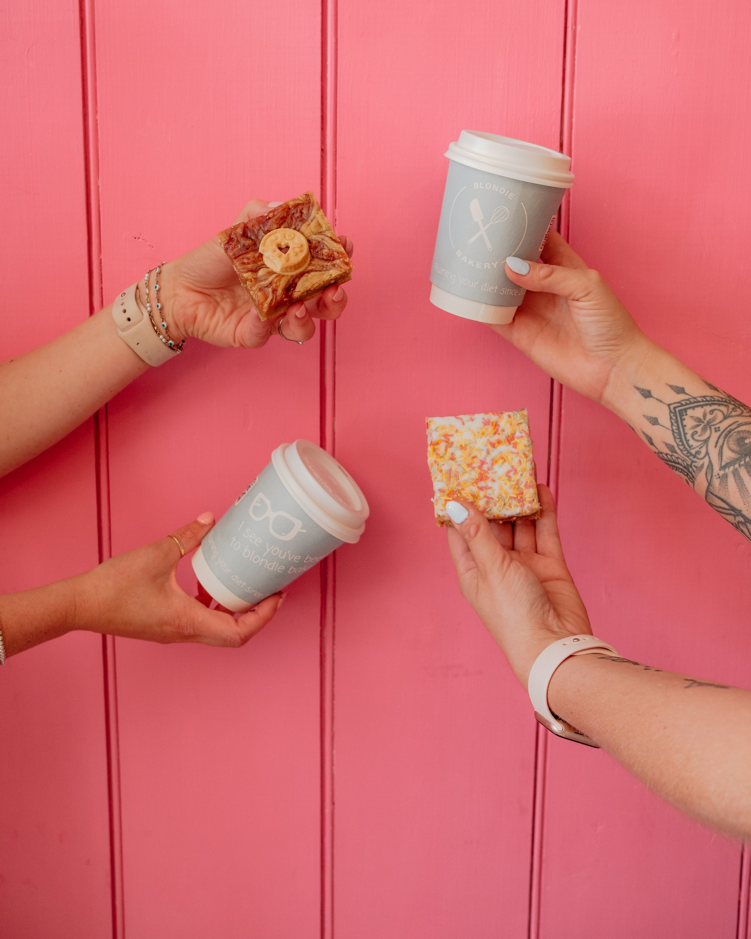 Three hands holding different snacks and coffee cups against a pink wooden background.