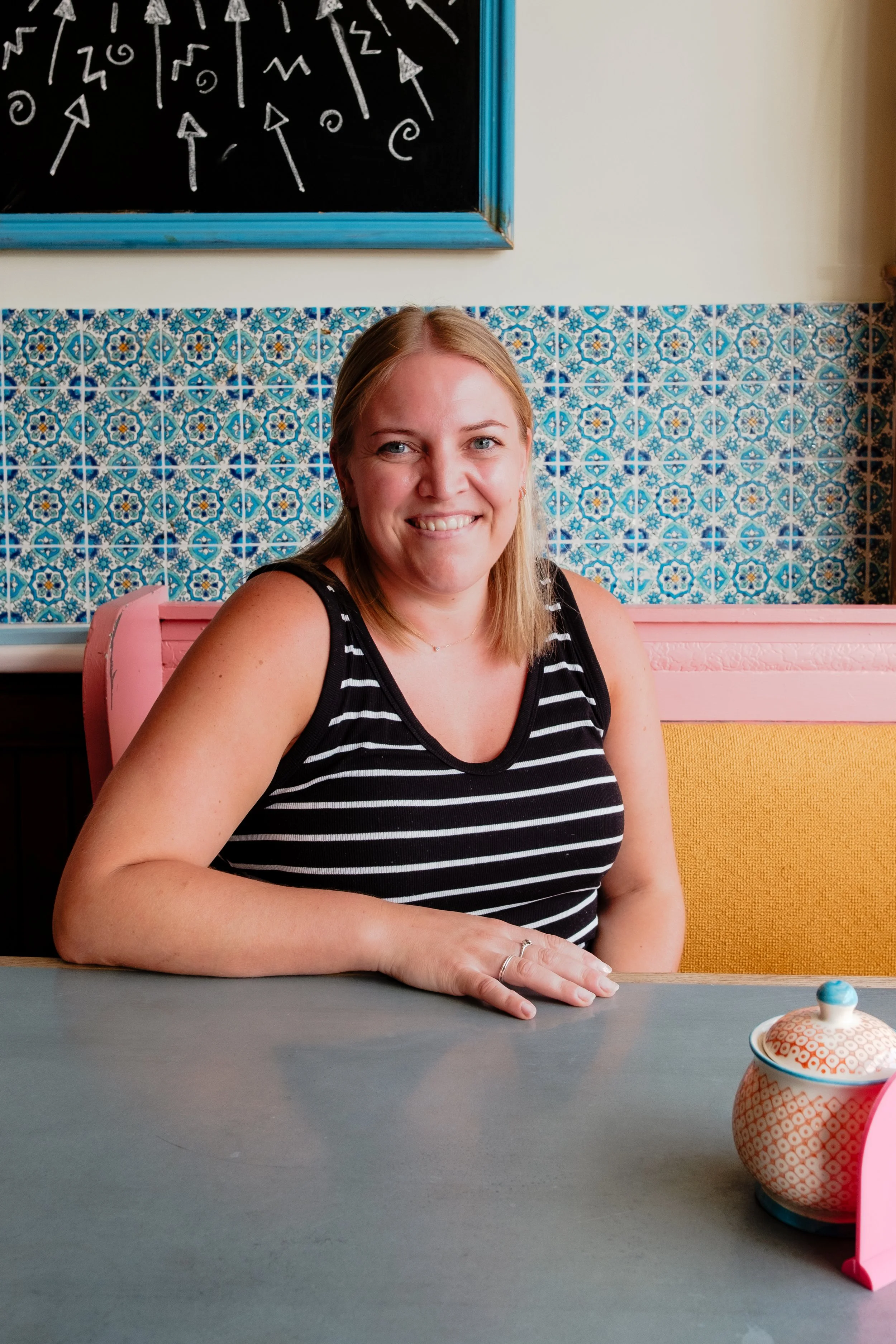 A smiling woman with blonde hair wearing a black and white striped sleeveless top, sitting at a grey table with a ceramic teapot in front of her. She is sitting against a background of blue patterned tiles and a chalkboard with drawn arrows and symbo