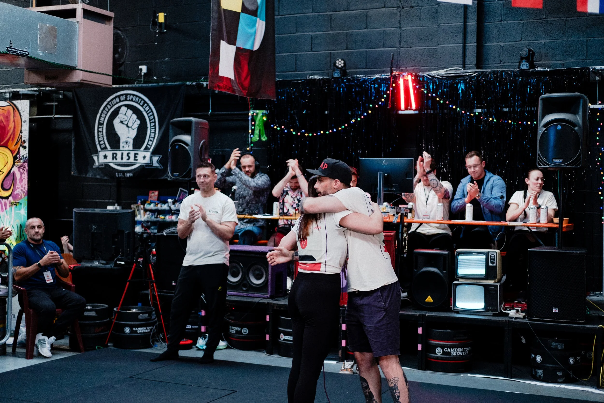 People are gathered in a room with black walls decorated with colorful string lights and a flag. Some individuals are dancing, while others are watching and clapping, with various equipment and speakers around them.
