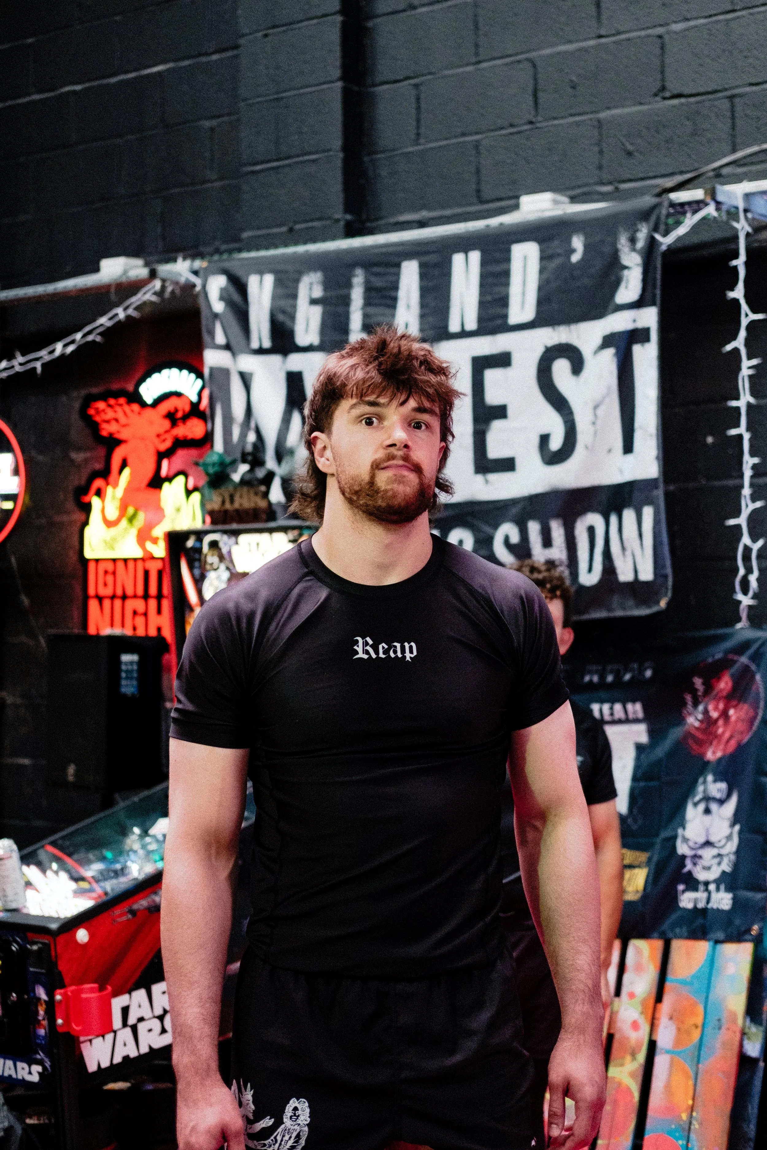 A man with brown hair and a beard wearing a black T-shirt with the word 'Reap' standing inside a store with dark walls, various neon signs, and posters including a Star Wars themed one in the background.
