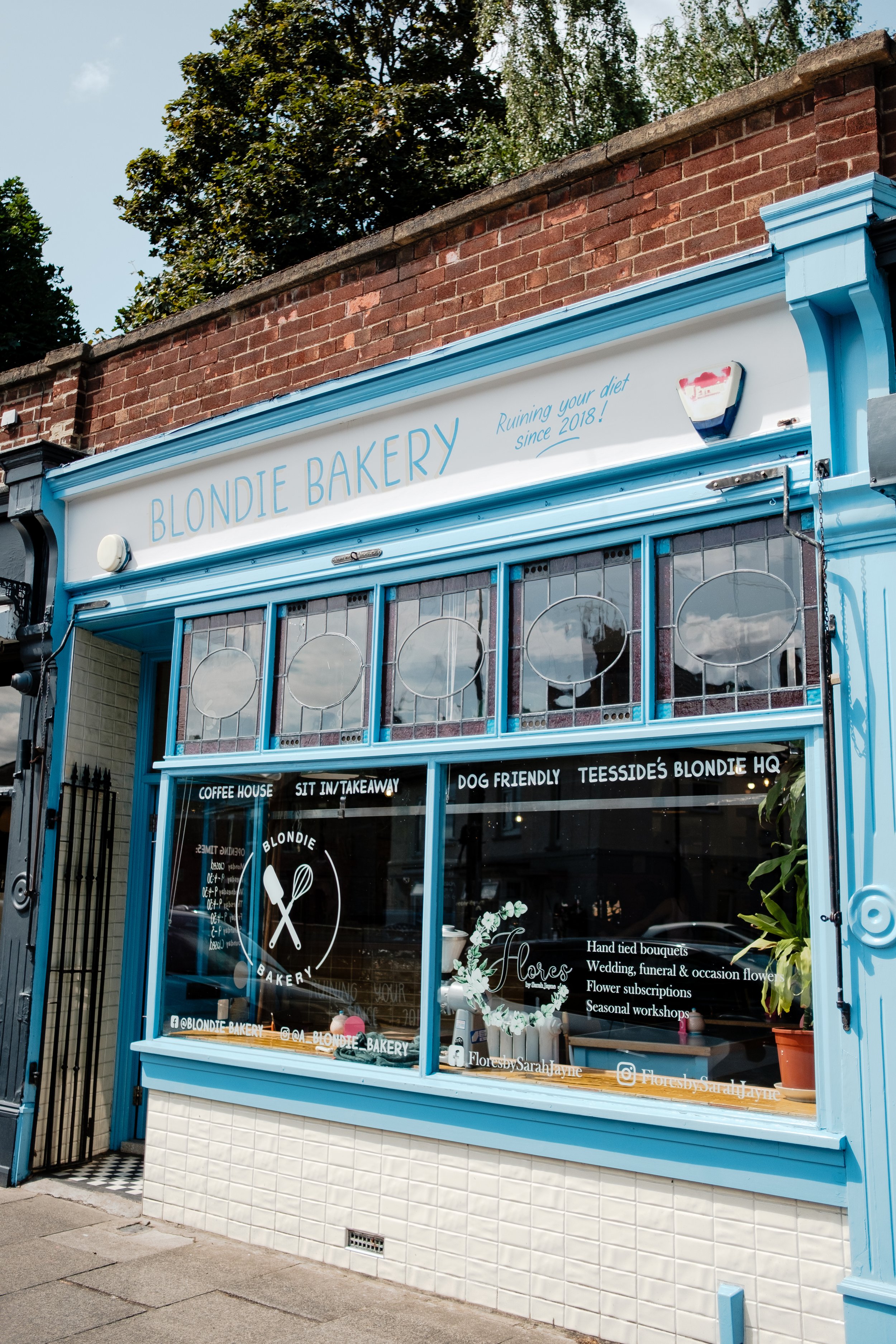 Exterior of a bakery called Blondie Bakery with a blue and white storefront, large window displaying bakery items, and a sign mentioning items like coffee, baked goods, and floral arrangements. There are trees and a brick wall behind the bakery.
